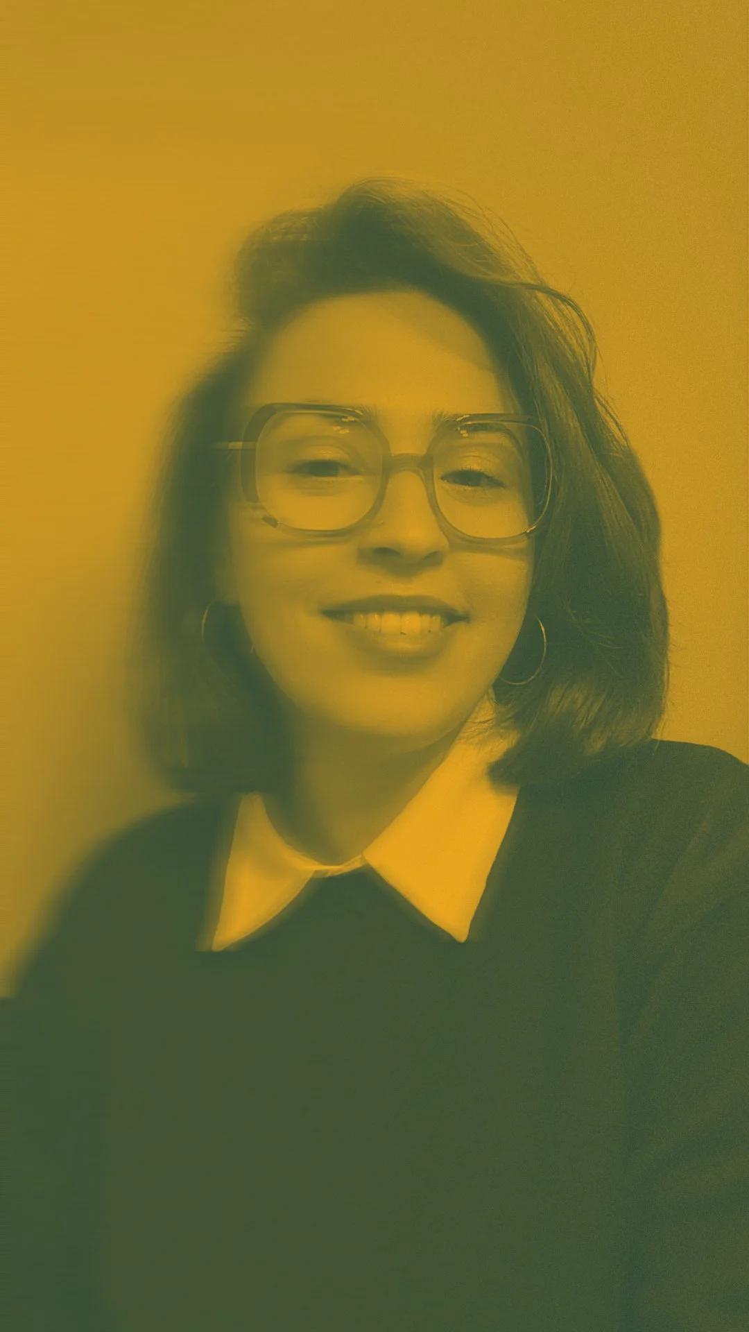 Portrait d'une femme souriante portant des lunettes, une chemise blanche sous un pull noir, avec des boucles d'oreilles, photographiée contre un mur beige.