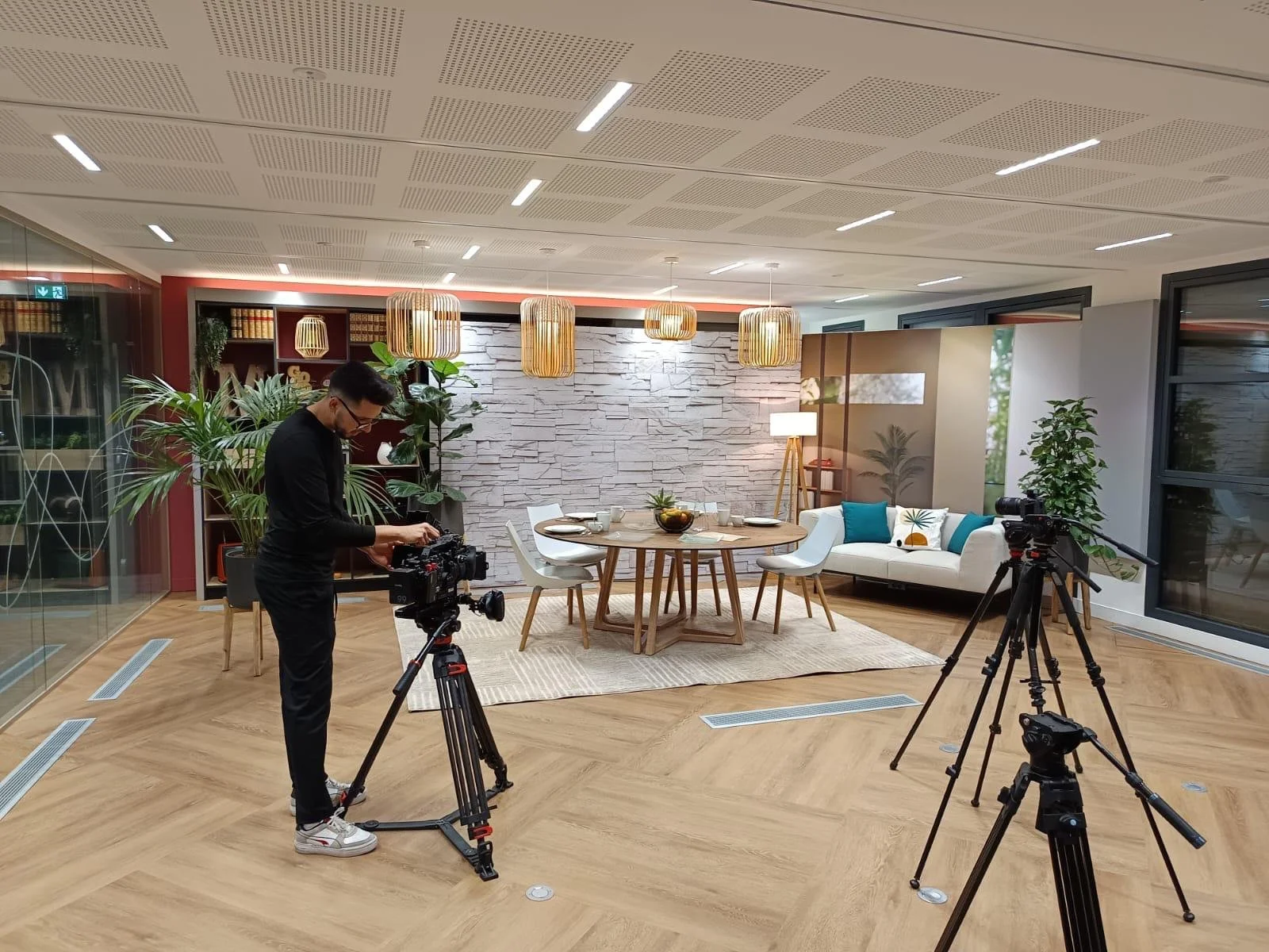 Studio de tournage avec un décor de salon comprenant un tapis, une table ronde, des chaises, un canapé blanc avec des coussins, des plantes vertes, et des lampes suspendues en rotin, avec un technicien en noir réglant une caméra.