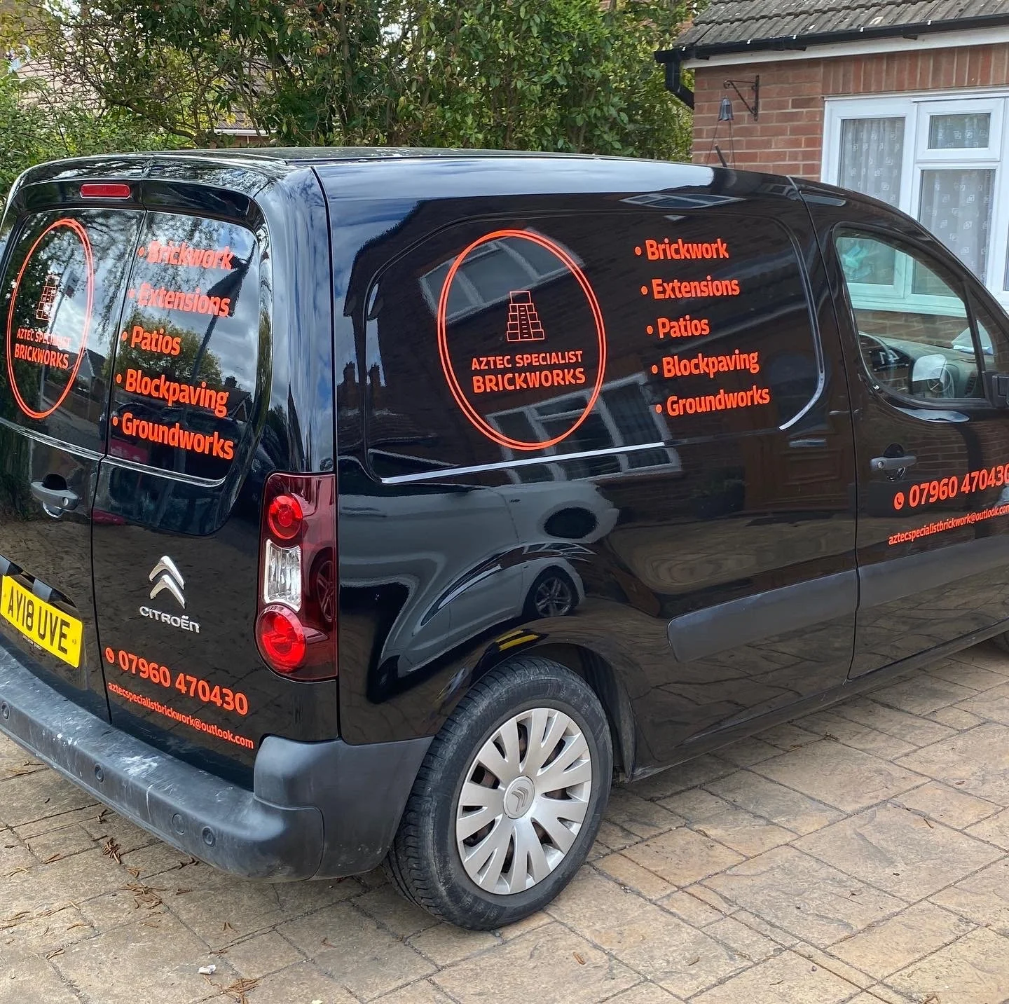 Black van with red lettering and logo for AZTEC SPECIALIST BRICKWORK, listing services such as brickwork, extensions, patios, block paving, and groundworks, parked on a brick driveway in front of a house with a brick exterior and window.