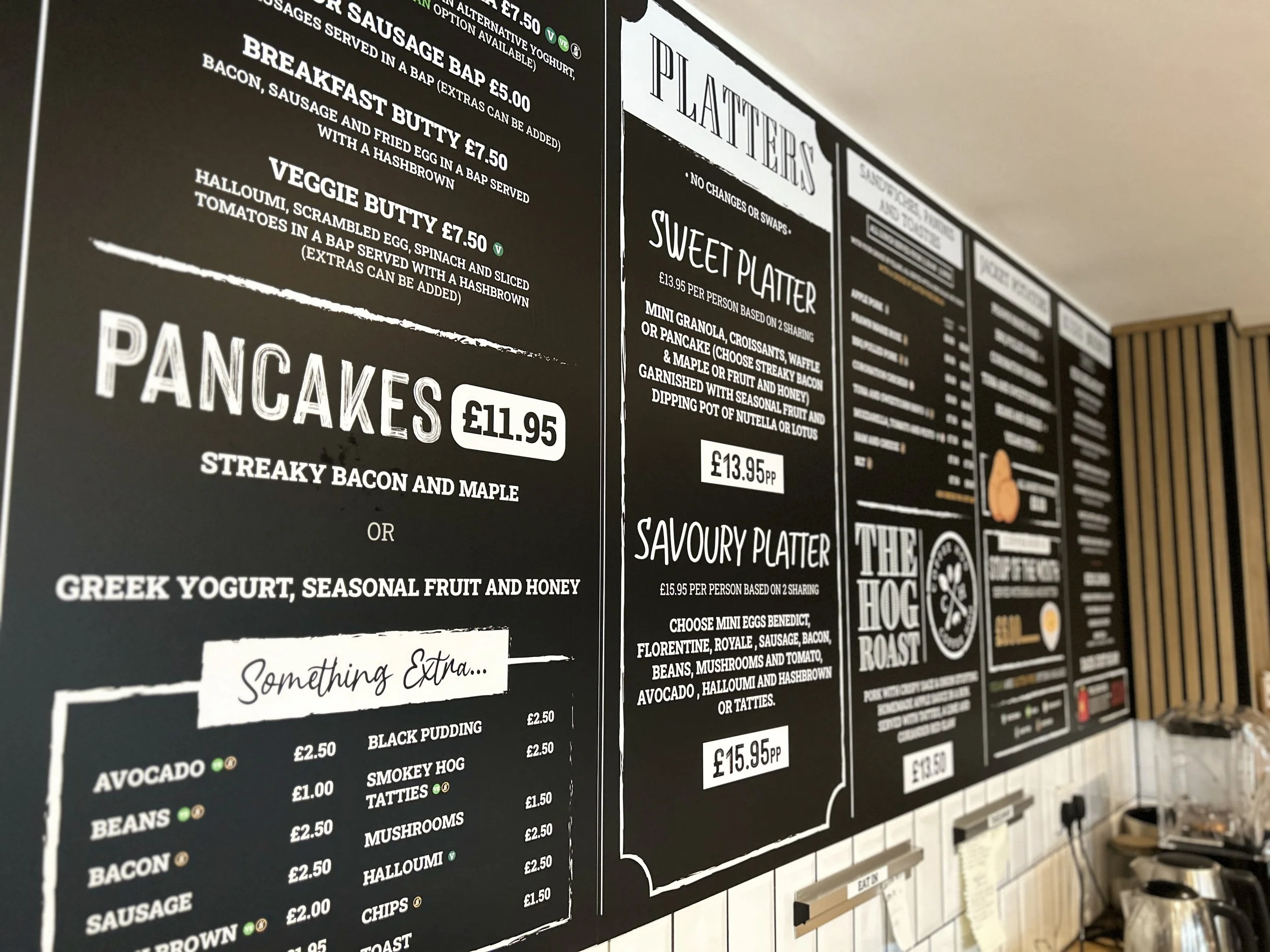 Close-up of a black menu board in a restaurant displaying breakfast, pancakes, and platter options with prices, alongside a section for extras and a small part of the cafe interior visible.
