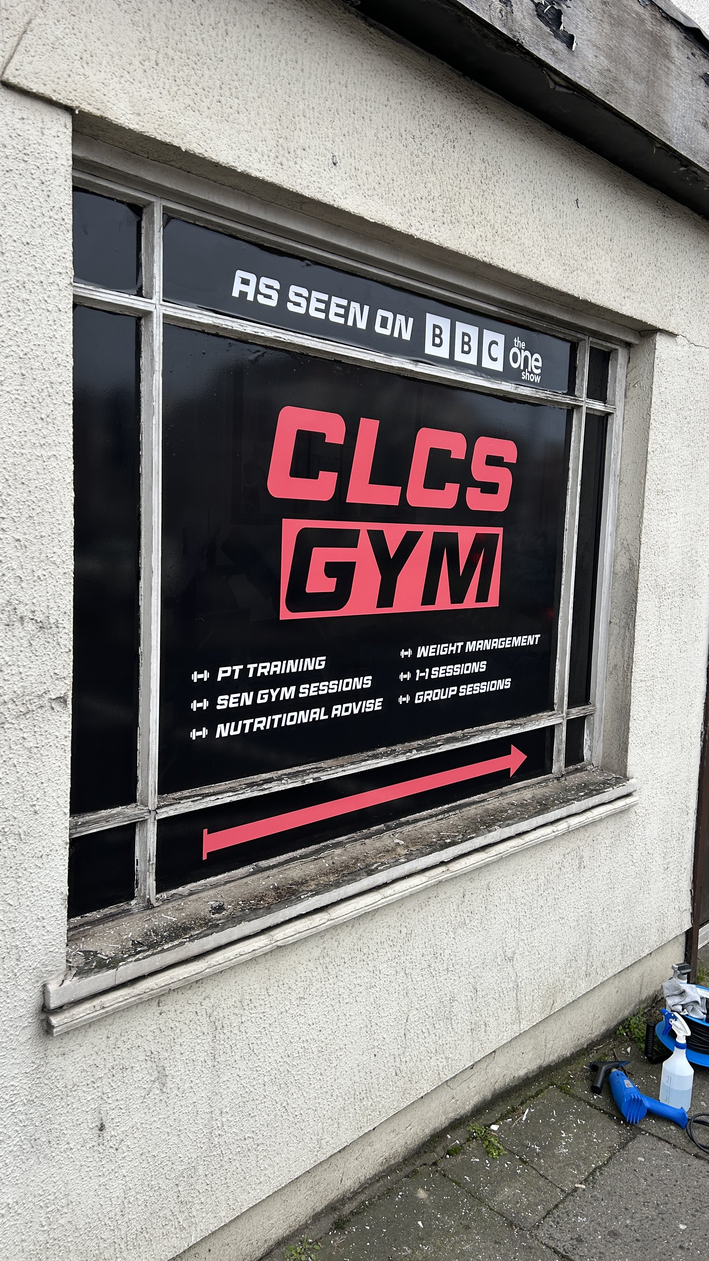 Sign for CLCS GYM on a building window with services listed, including PT training, SEN gym sessions, nutritional advice, weight management, 1-1 sessions, and group sessions, with an arrow pointing to the right.