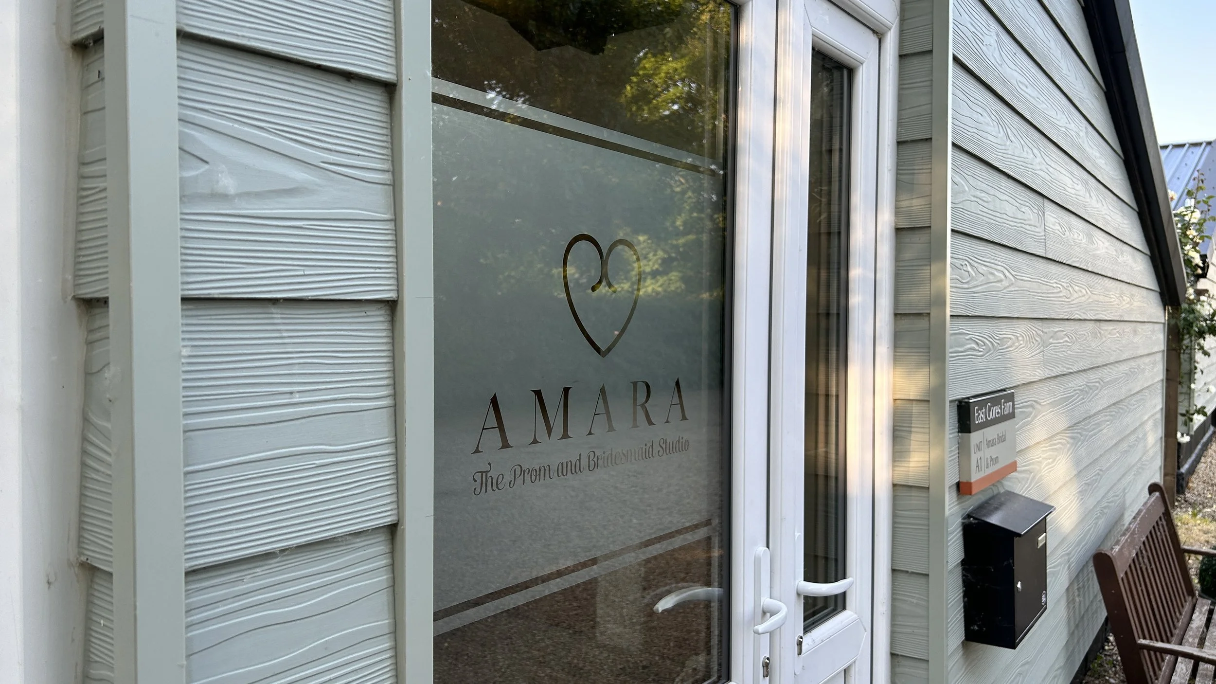 Glass door with a logo and text for Amara, a prom and bridemade studio, on a white exterior building with siding. A black mailbox and a wooden bench are outside.