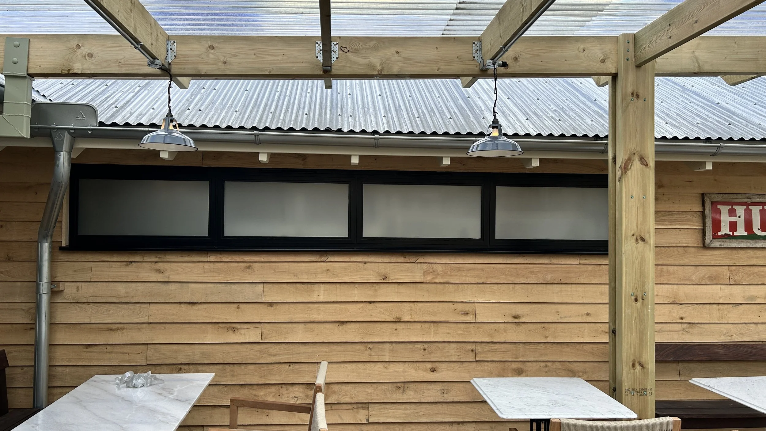 Wooden exterior of a building with a large horizontal window, metal roof, hanging metal light fixtures, and outgoing tables.