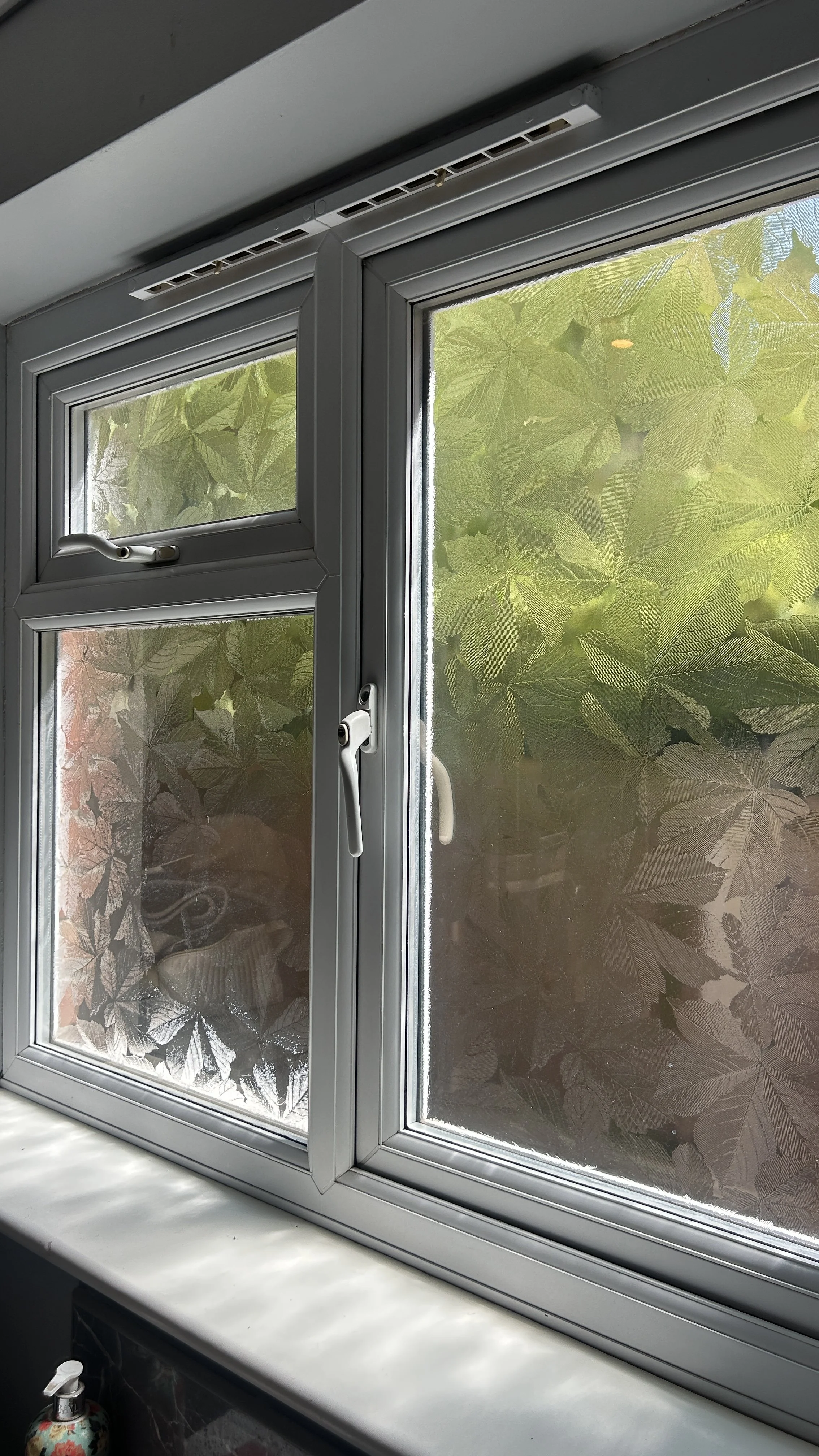 A white-framed window with frosted glass featuring a leaf pattern. The window has a handle on the right side, and the top part of the frame has a vent with multiple slots. Outside the window, green leaves are visible.