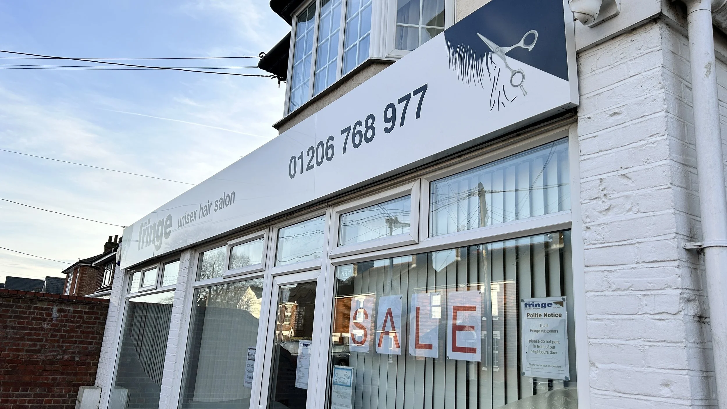 Exterior of a unisex hair salon called 'The Fringe' with a large sale sign in the window and a polite notice from the salon, located in a residential neighborhood with a brick wall and neighboring houses visible.