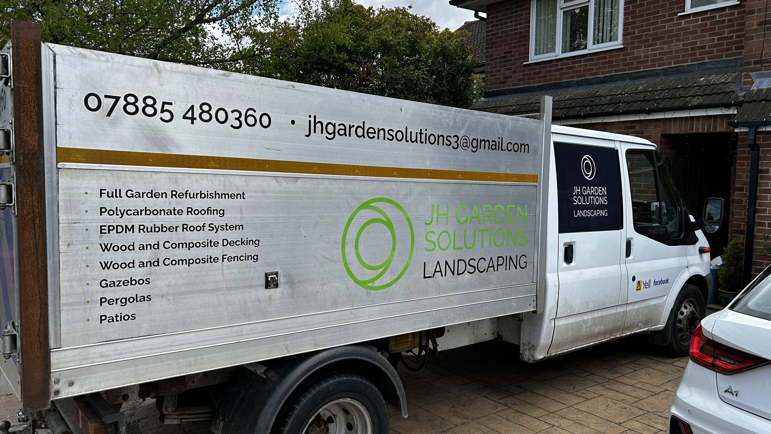 A white work truck with a sign for JH Garden Solutions Landscaping parked in a driveway next to a brick house. The sign lists services such as full garden refurbishment, polycarbonate roofing, EPDM rubber roof system, wood and composite decking, fenc