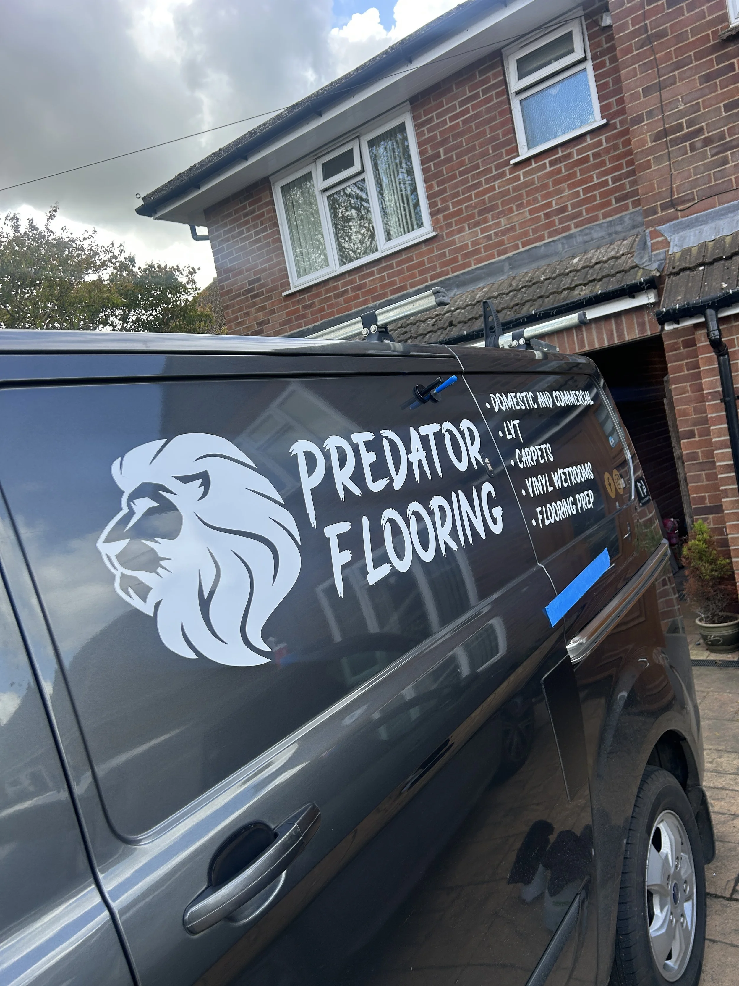 A vehicle with a business logo related to flooring services parked in front of a brick house with a window. The logo includes a lion's head and lists services like domestic and commercial flooring, LVH, carpets, vinyl, wetrooms, and flooring prep.