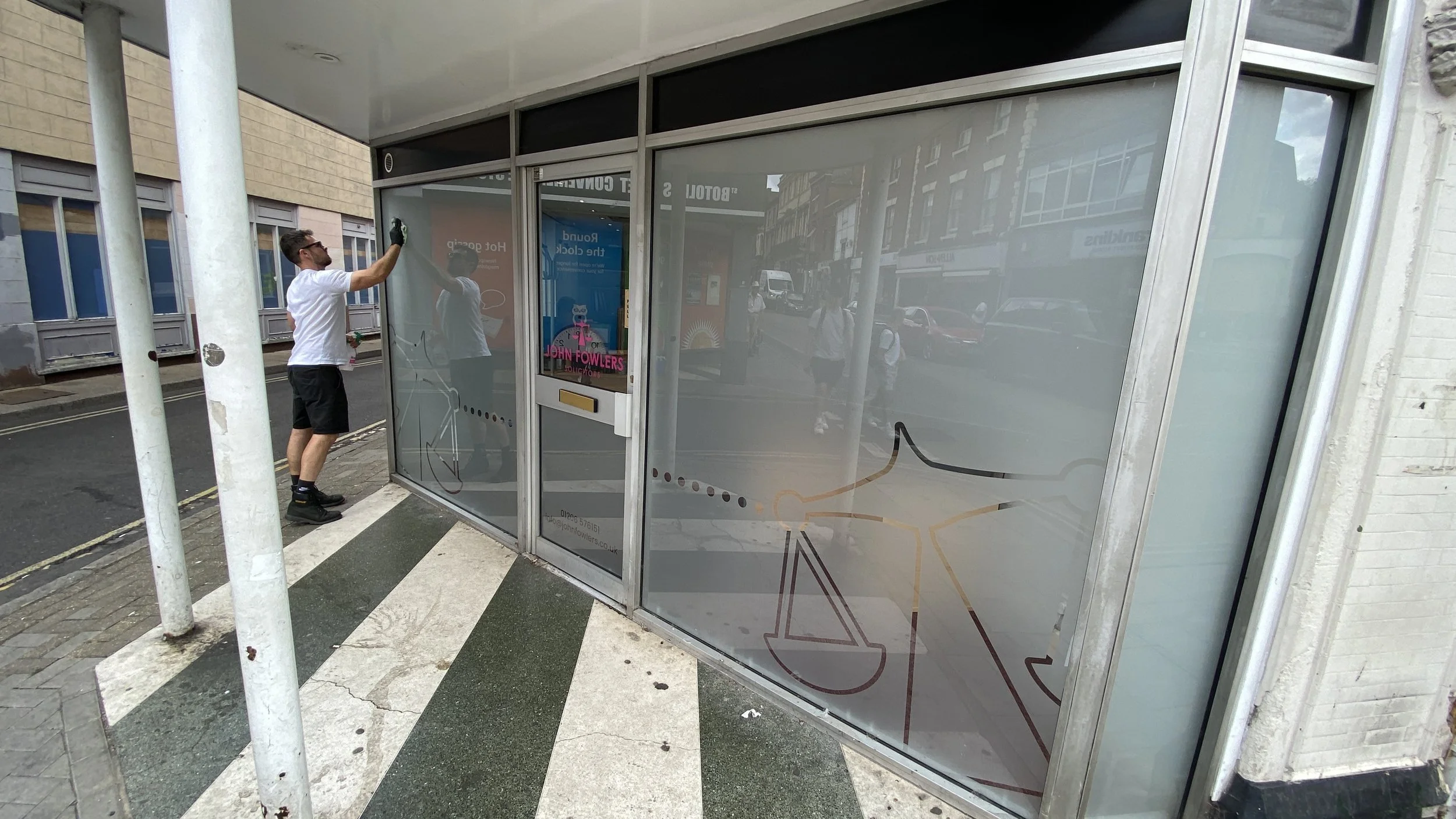 A man cleaning the glass window of a storefront on a city street. The storefront has a large glass panel with some colorful designs and text. The man is wearing a white T-shirt, black shorts, and gloves, and is standing on the sidewalk.