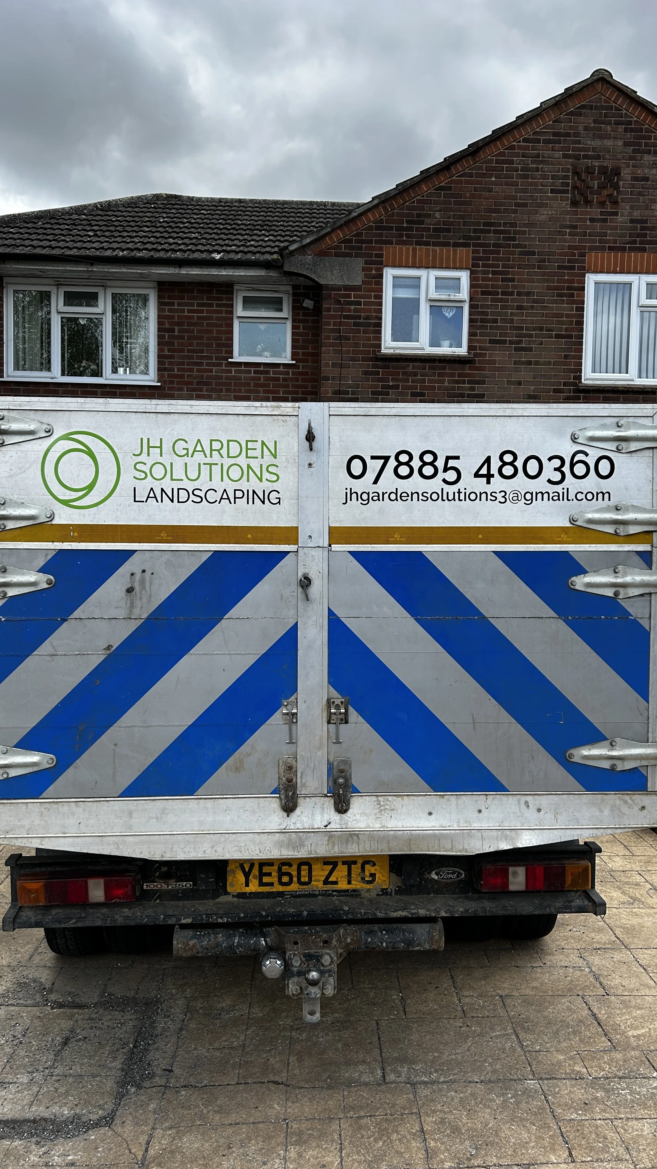 The back of a white utility vehicle with a sign for JH Garden Solutions Landscaping, including a phone number and email, parked in front of a brick house with white-framed windows and a dark tile roof.