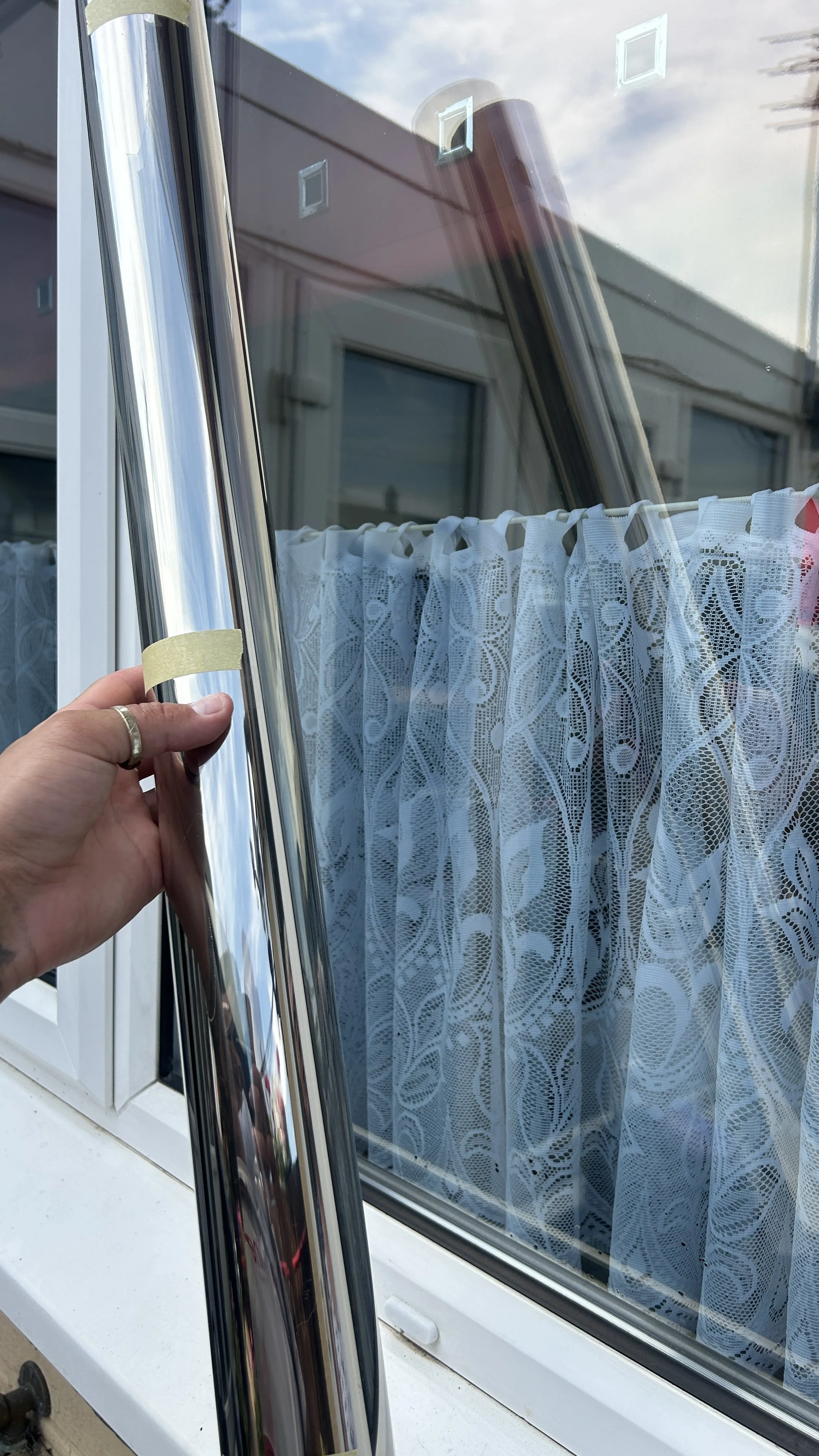 Person holding a metal frame window shows a lace curtain inside a house. The window is open, with reflections of a cloudy sky in the glass.