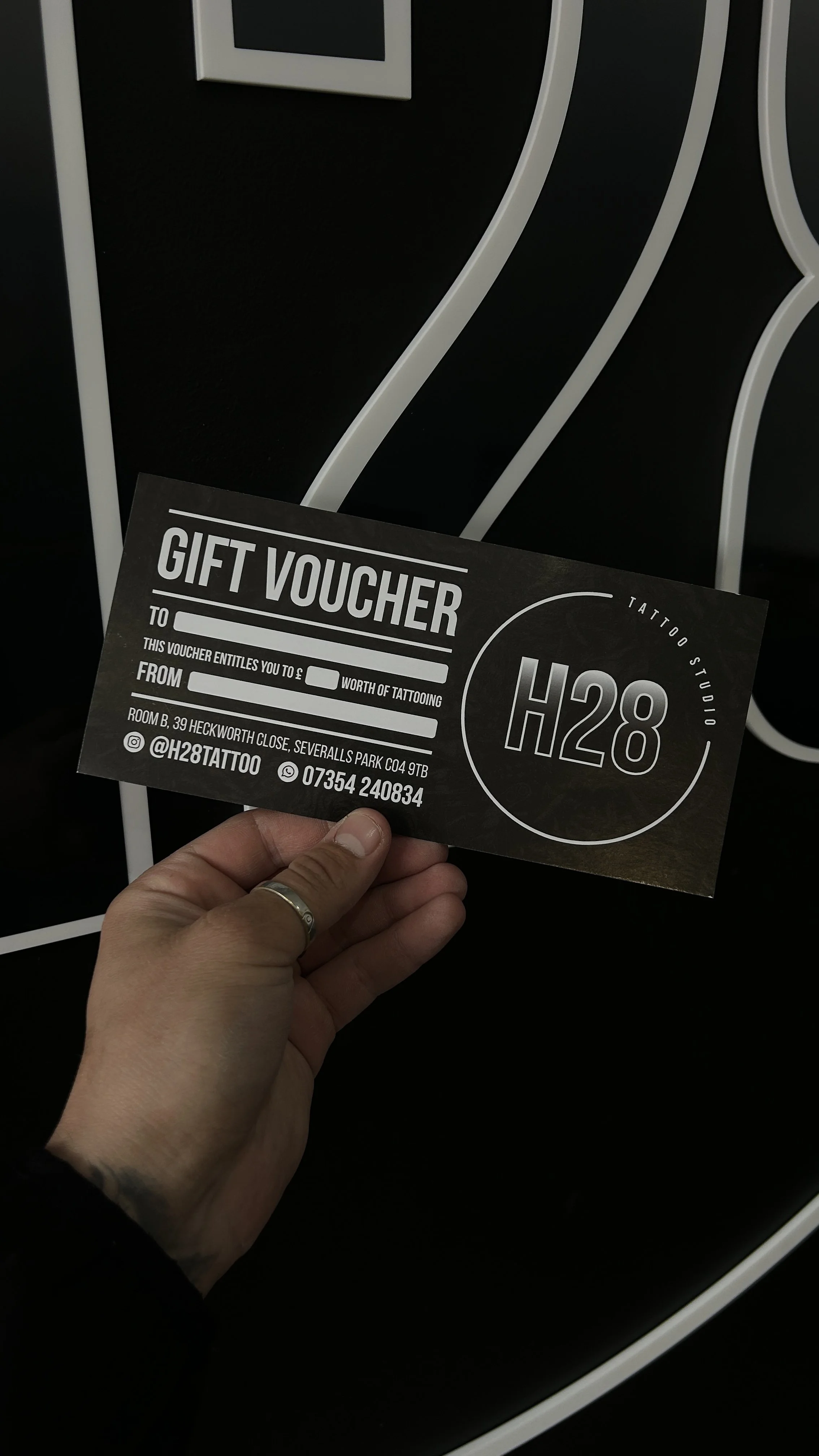 A person holding a black gift voucher for a tattoo studio, with the studio's contact information and social media handle visible. The background features black and white abstract artwork.
