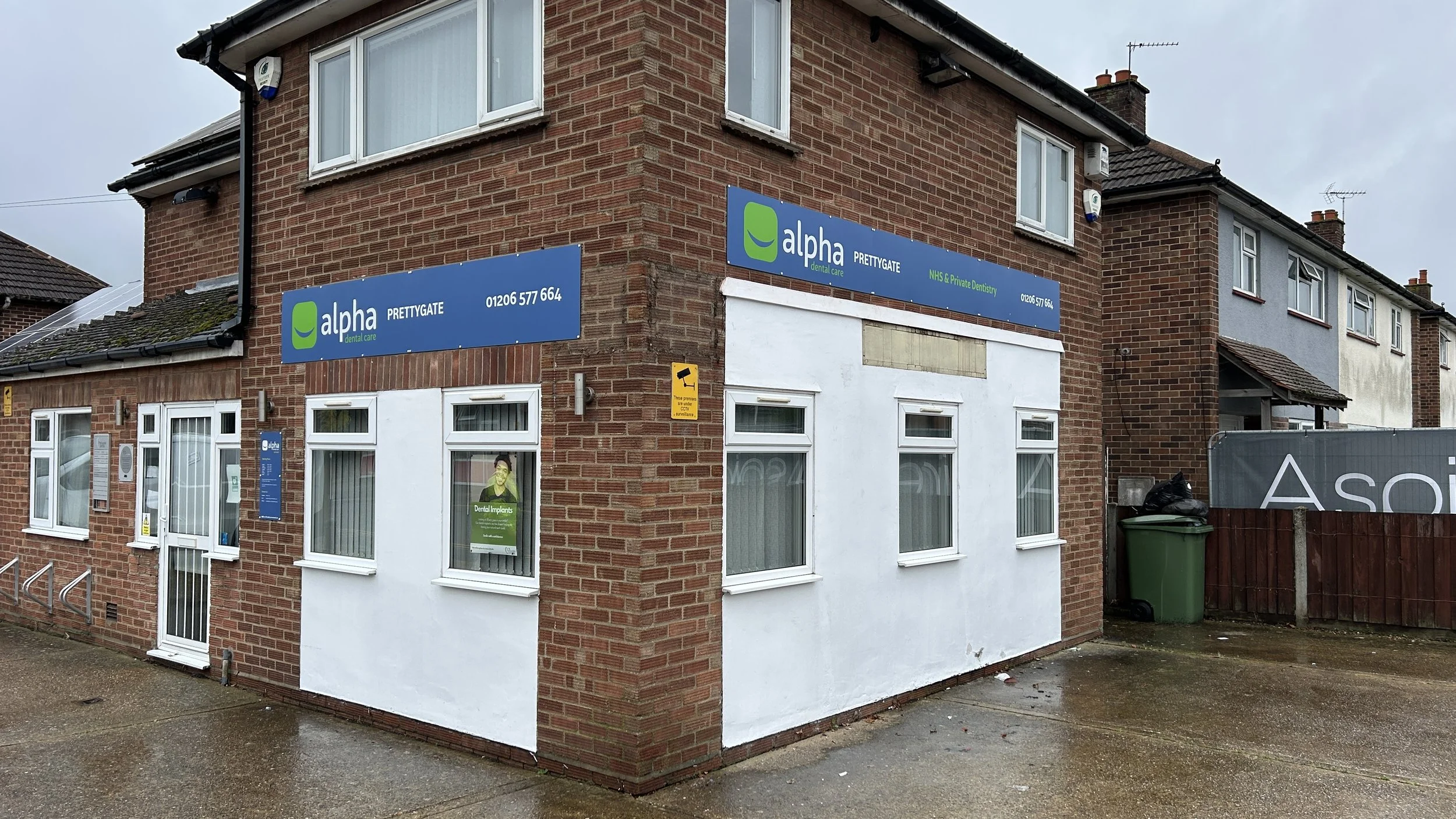Corner view of a brick building with dental care signs for Alpha Dental Care, showing upper and lower windows, a small poster in the window, a green trash bin outside, and a security camera on the corner.