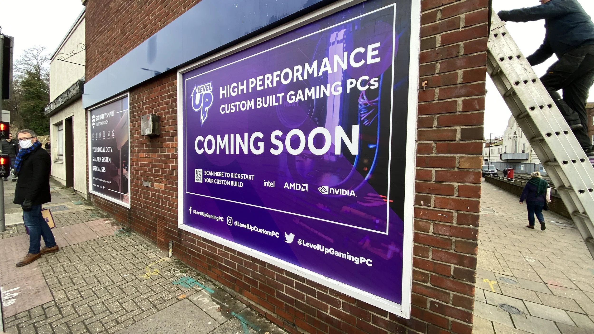 Large purple sign on a brick wall advertising a new high-performance custom gaming PC shop, coming soon. The sign includes logos of Intel, AMD, and NVIDIA, and prompts viewers to scan for more info. Nearby, a man with a face mask stands on the sidewa
