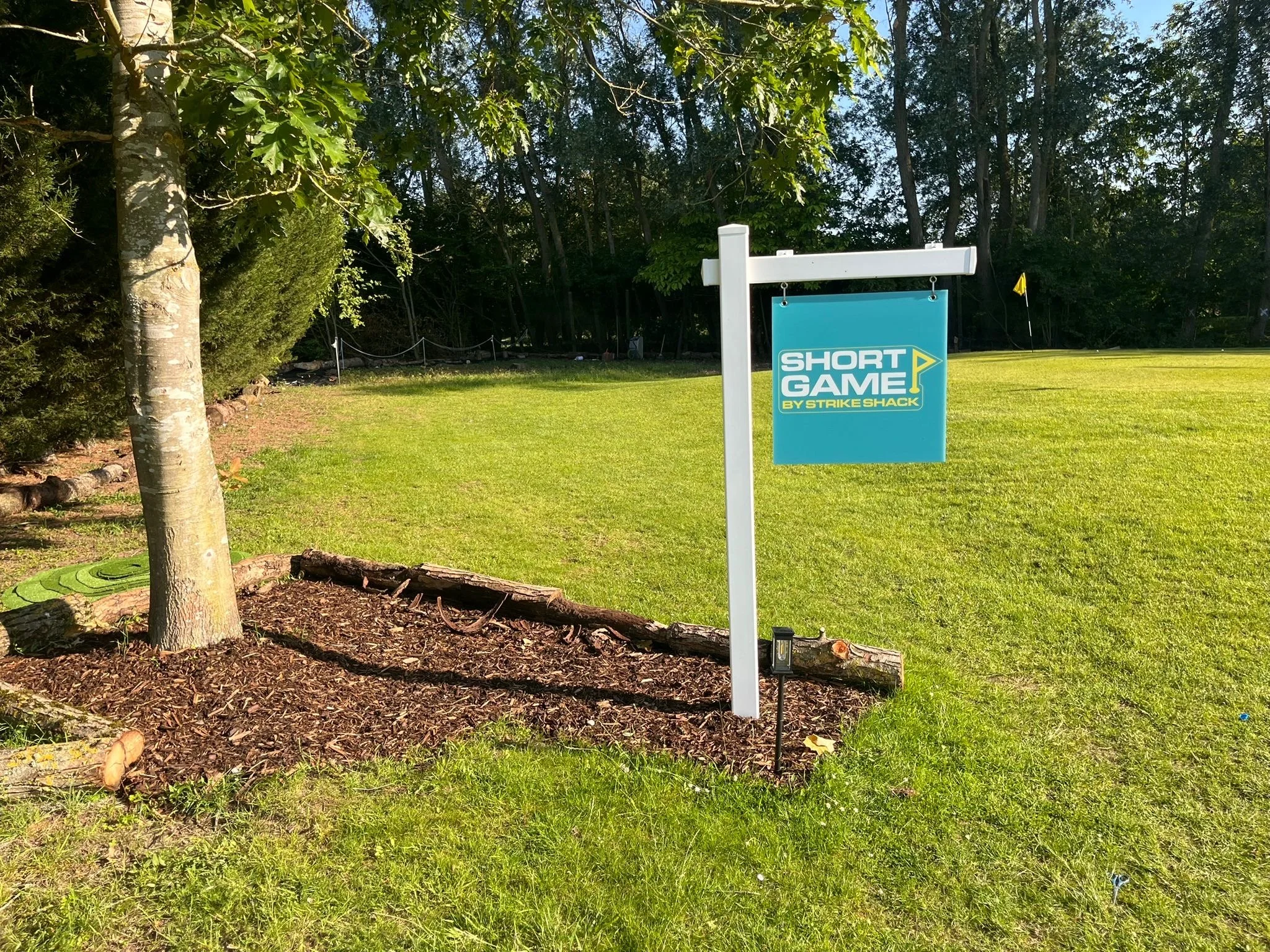 A small blue sign on a golf course with the words 'Short Game by Strike Shack' and a yellow flag icon. The background features green grass, trees, and a wooded area.