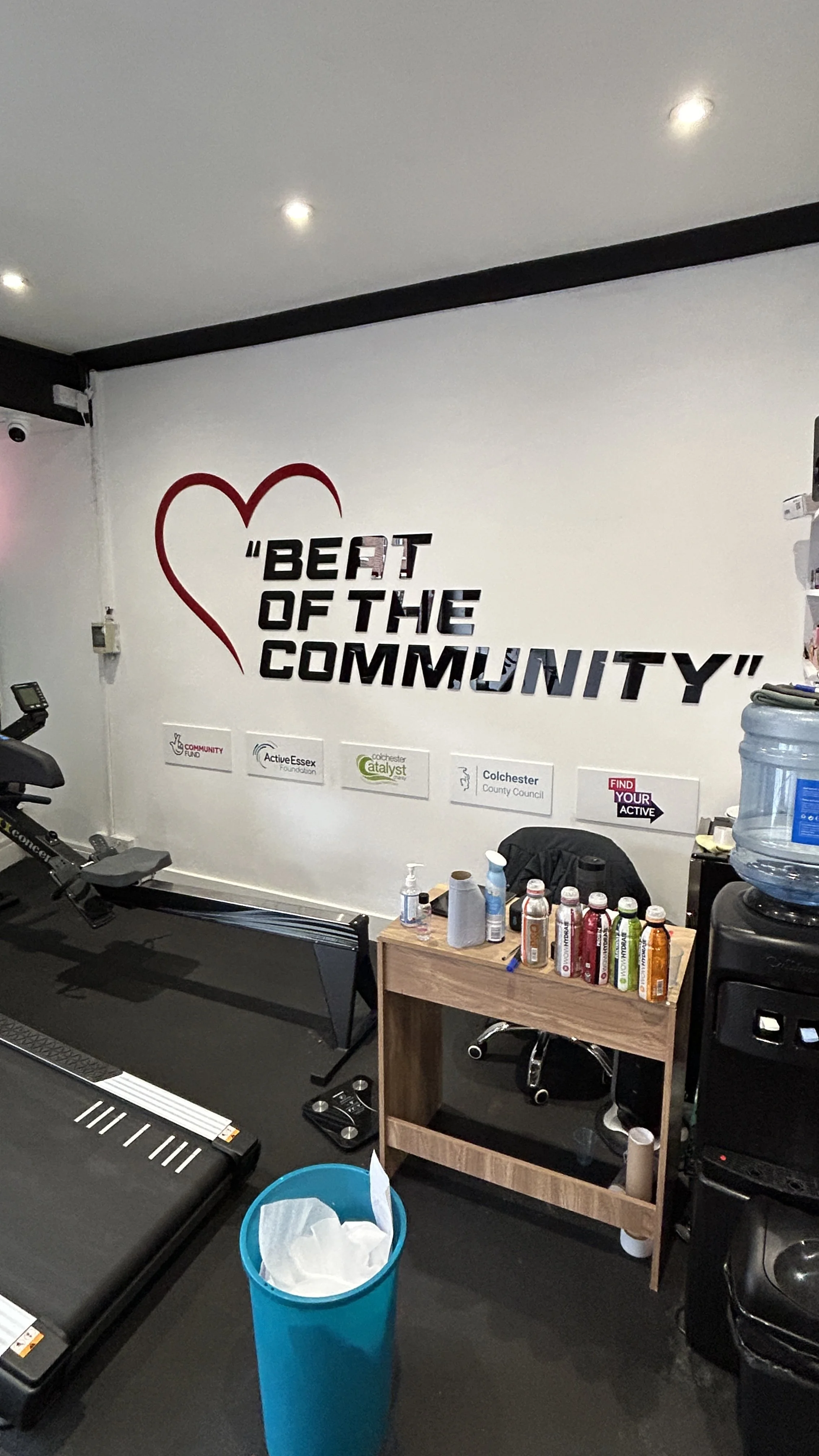 An exercise room with a rowing machine, small wooden table holding beverages and supplies, water cooler, trash bin, and a motivational wall mural that reads "Beat of the Community" with a heart outline and logos of local organizations.