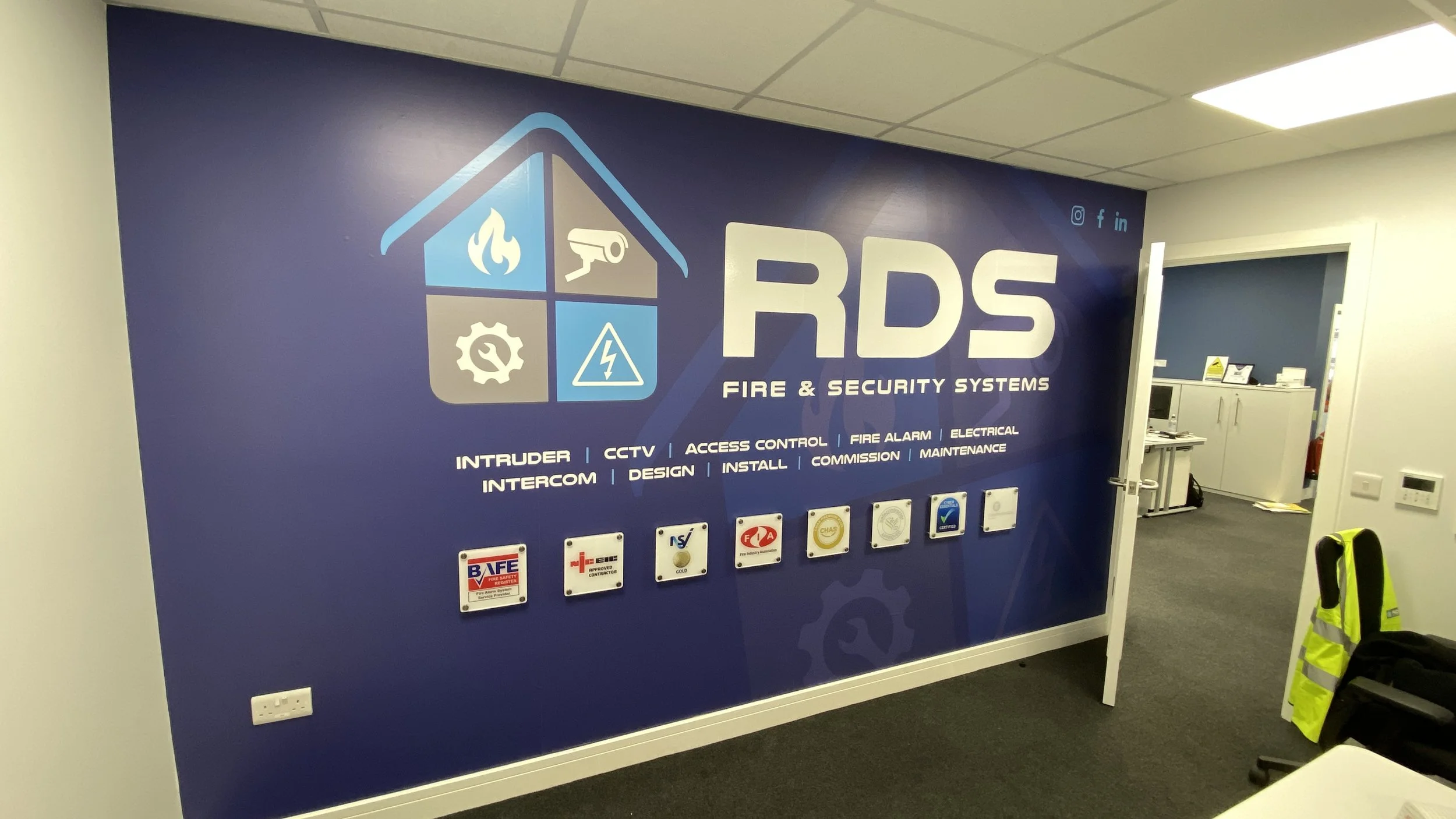 Blue wall with RDS Fire & Security Systems signage, icons for fire, security, CCTV, and electrical, certificates, and notices displayed beneath, in an office setting.