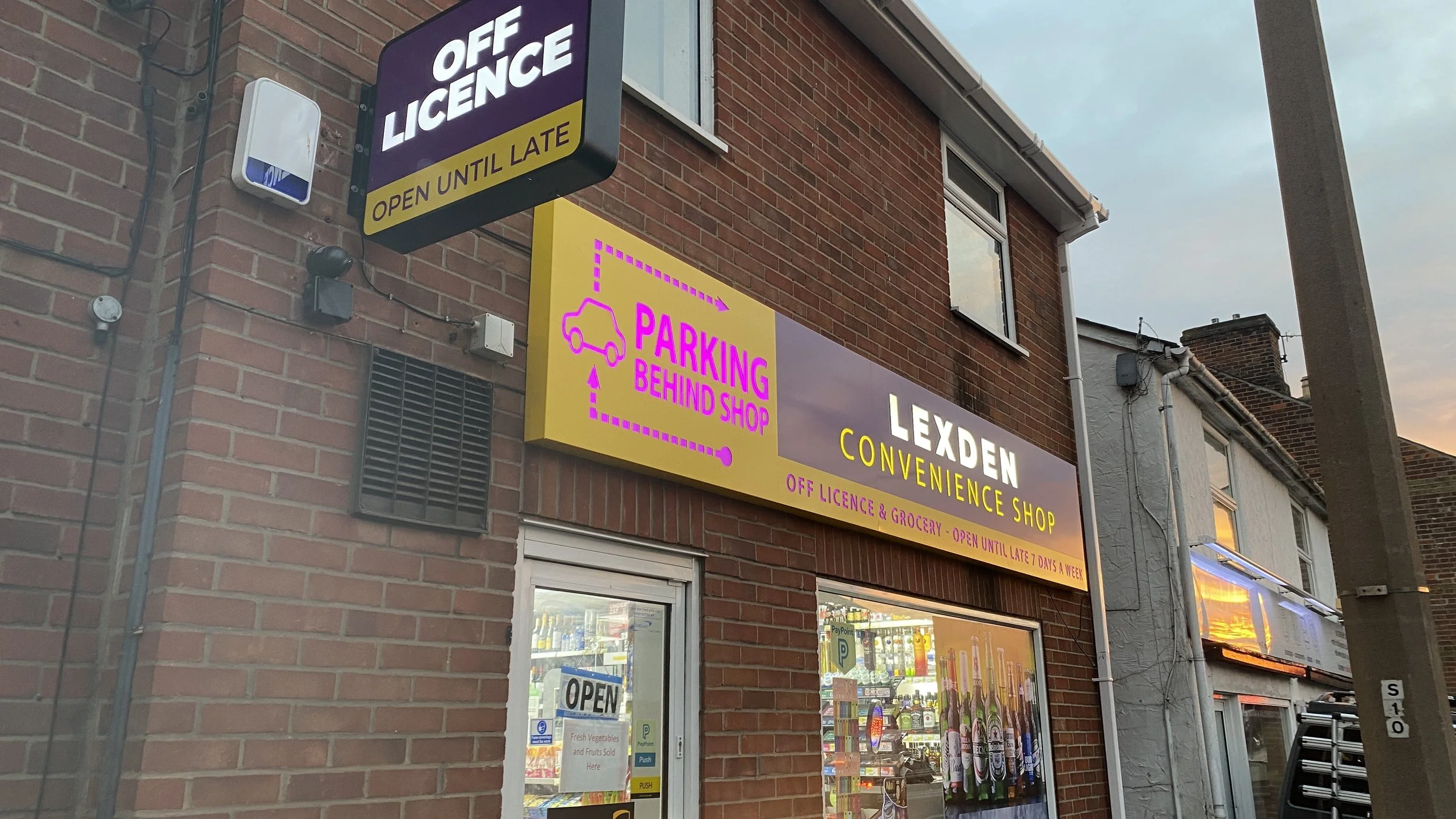 Signage for Lexden Convenience Shop showing parking instructions and shop hours, on a brick building exterior with a window and door, with sunset sky visible.