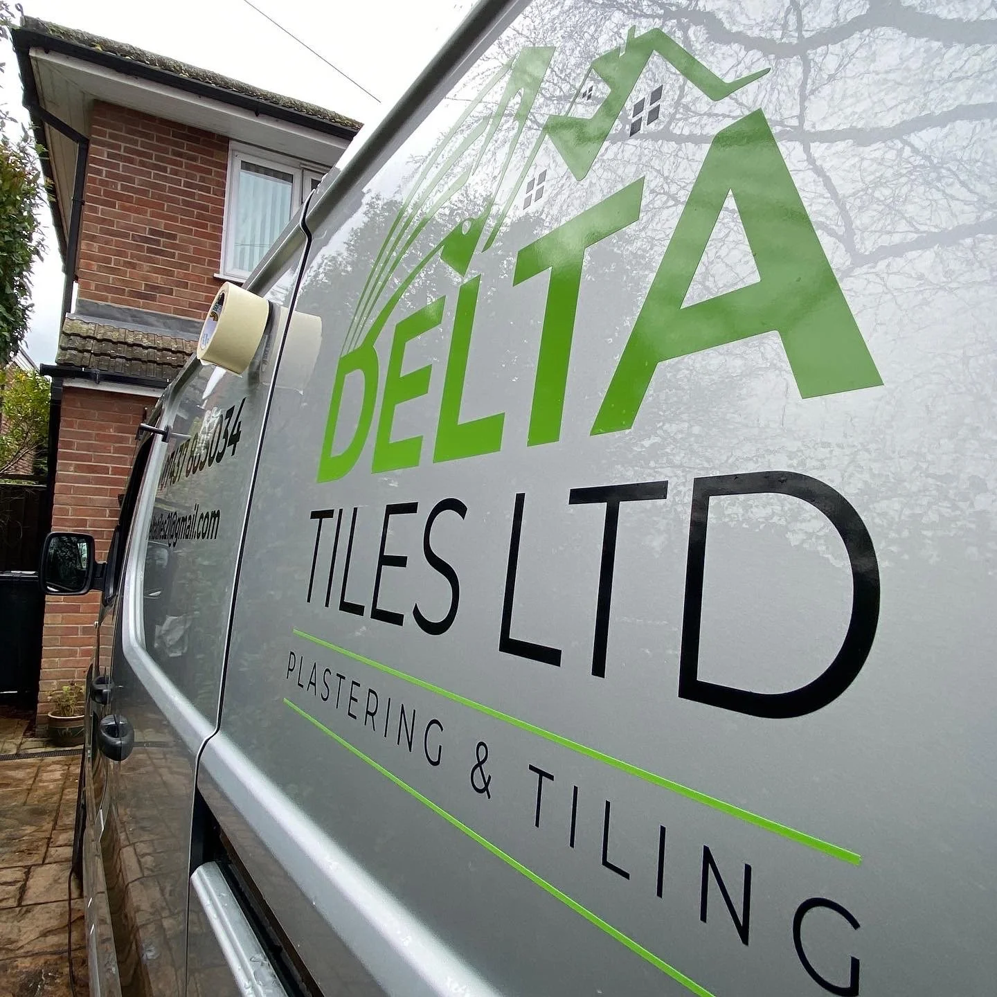 A vehicle with a sign advertising a company called Delta Tiles Ltd, which specializes in plastering and tiling services. The sign features green and black text on a white background. The photo is taken from the side of the vehicle, with a house and t