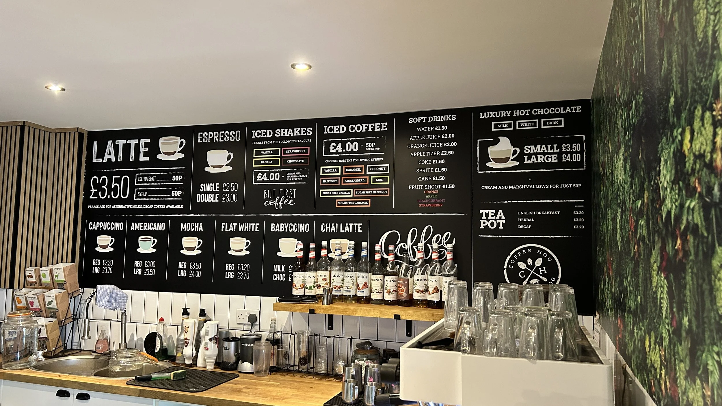 Coffee shop menu board with various drinks and prices, bottles of syrup on shelf, upside-down glasses, and a wooden counter with equipment and a partial view of green leaves wallpaper.