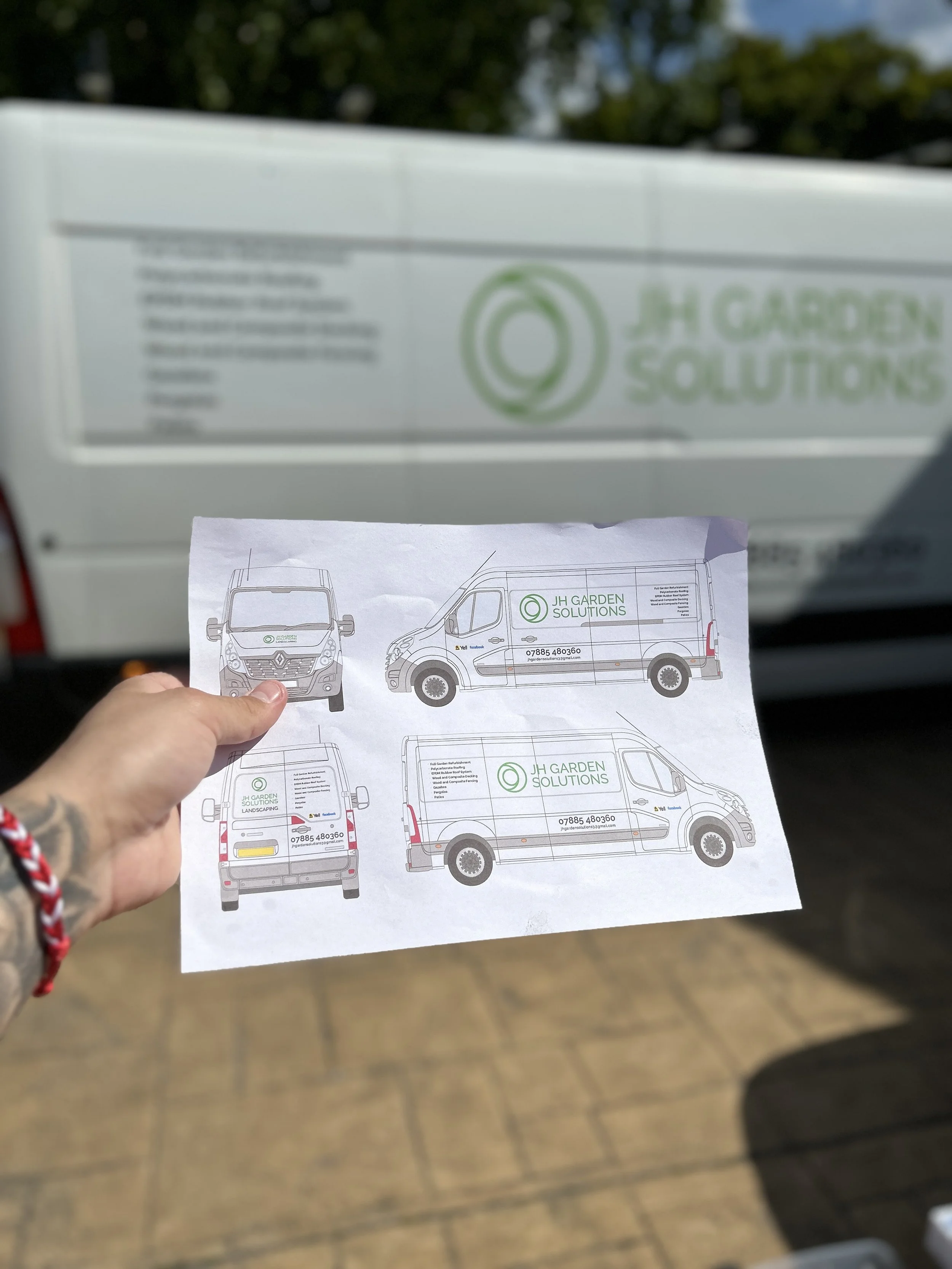 Hand holding a printed delivery van design sketch in front of a real van with matching branding, outdoor setting.