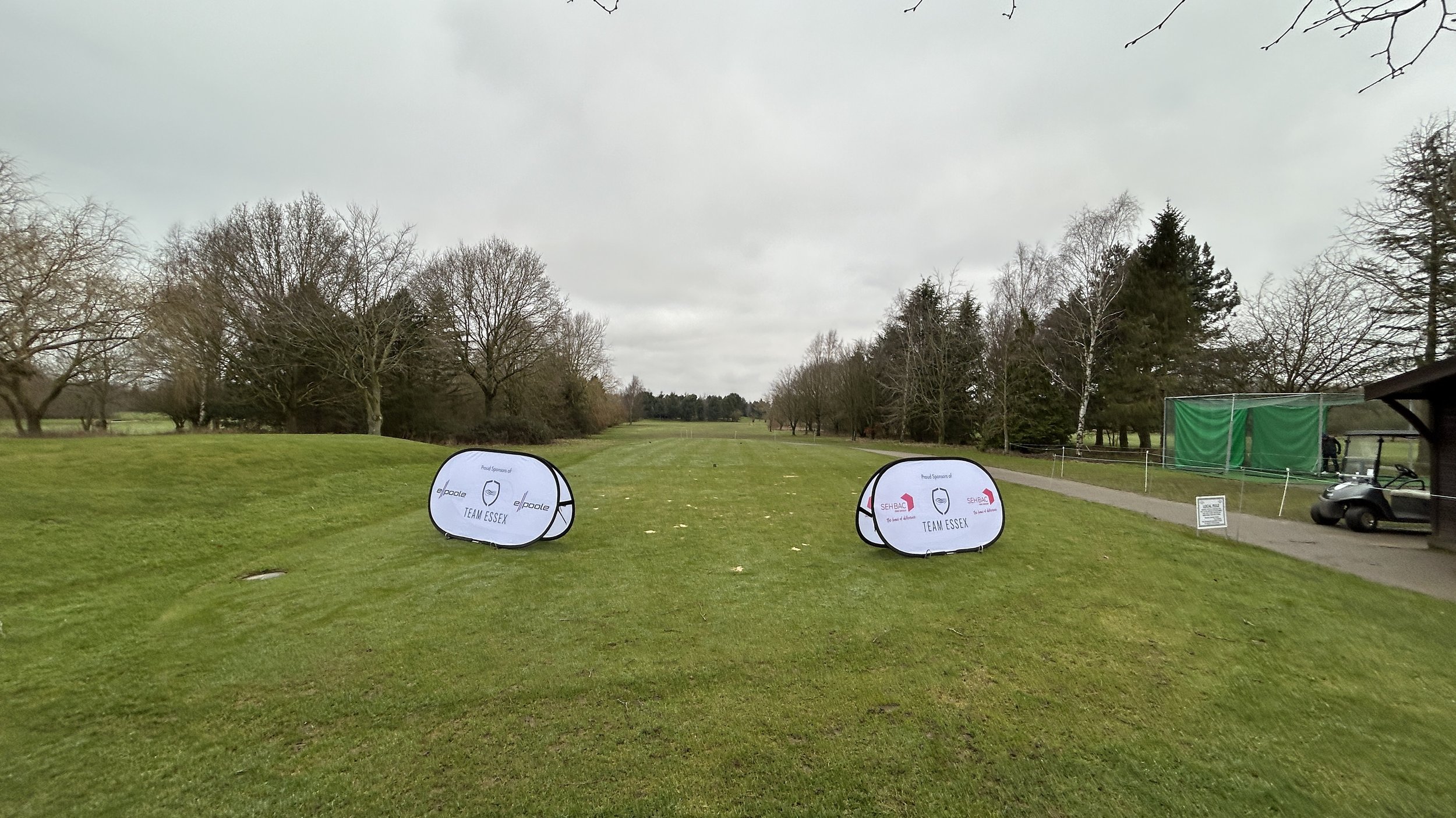 A view of a golf course with a cloudy sky, some leafless trees, and two promotional banners for Team Essex on the grass.