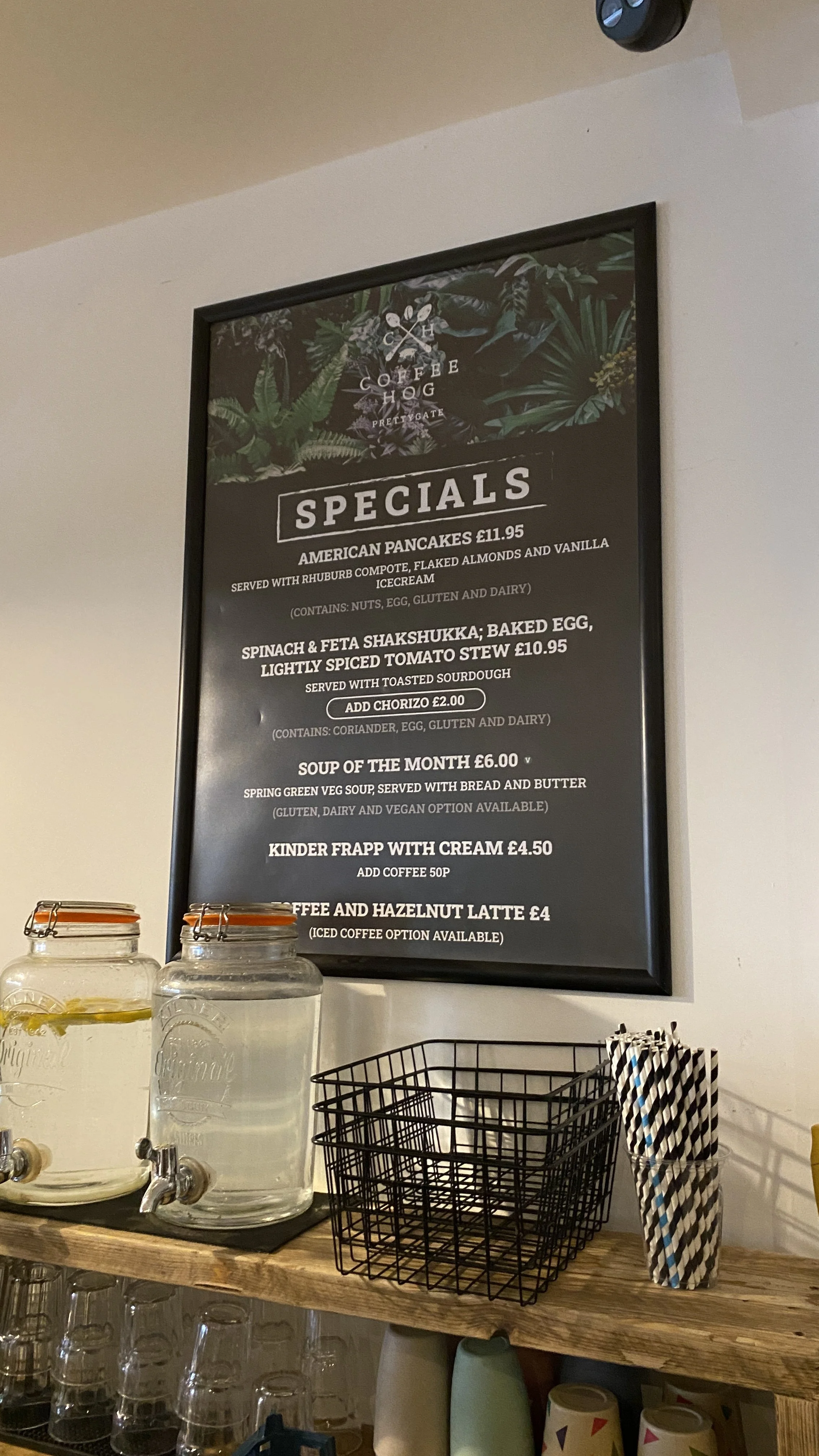 A blackboard menu with drinks and food specials at a cafe, including American pancakes, shakshuka, soup of the month, Kinder frappe, and coffee options, above a wooden counter with glass jars and striped paper straws.