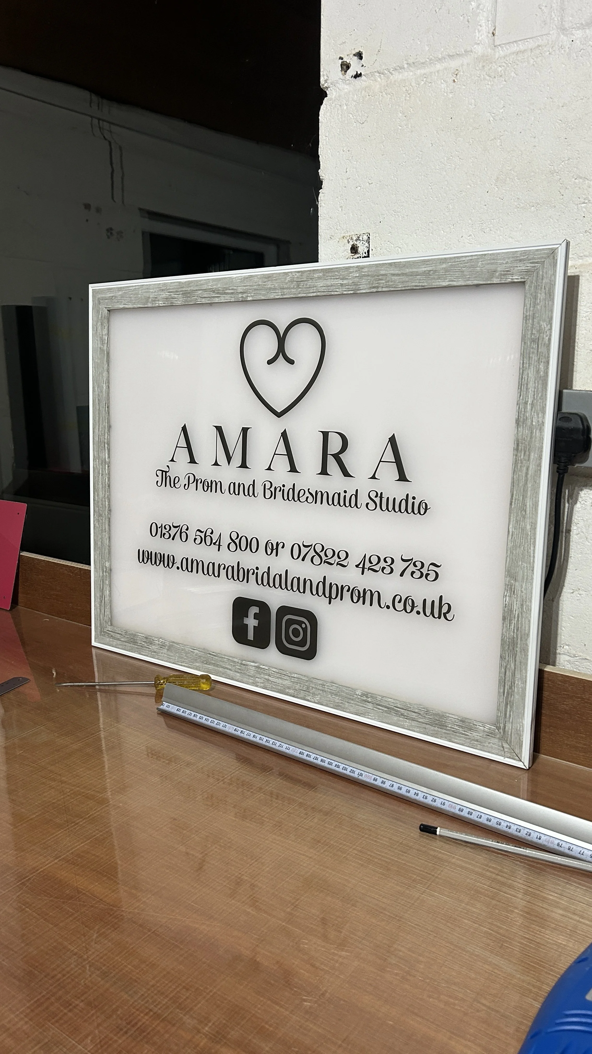 A white sign with a heart logo and text that reads 'AMARA The Prom and Bridesmaid Studio' along with contact information and social media icons for Facebook and Instagram, placed on a wooden table.