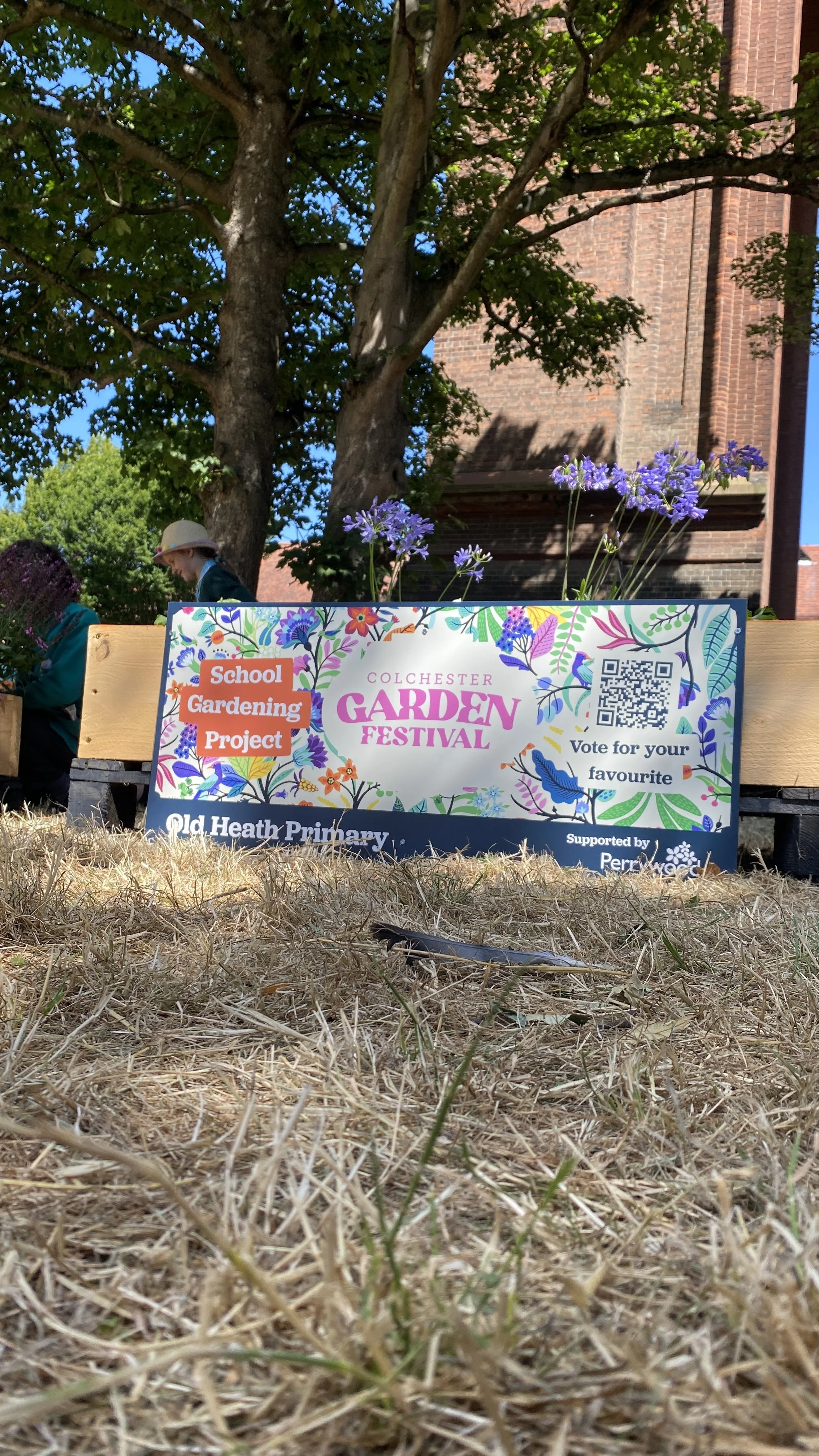 Sign for Colchester Garden Festival promoting school gardening project supported by Perrywood, with purple flowers and trees in the background.