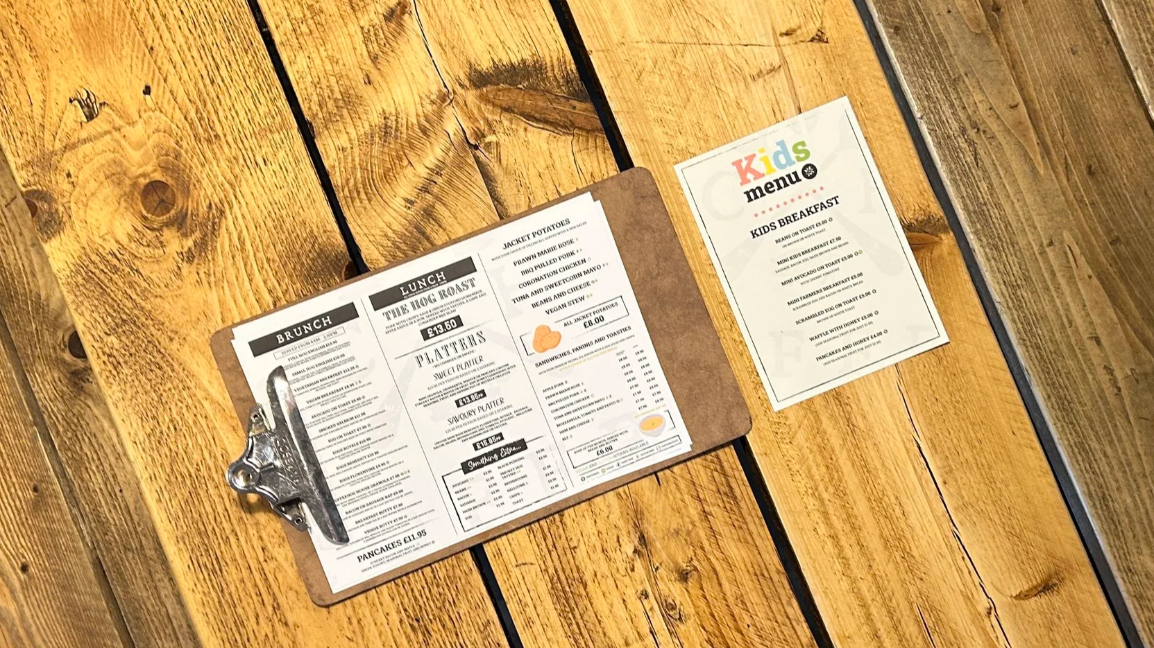 A wooden table holding a brunch menu on a clipboard and a Kids menu flyer.
