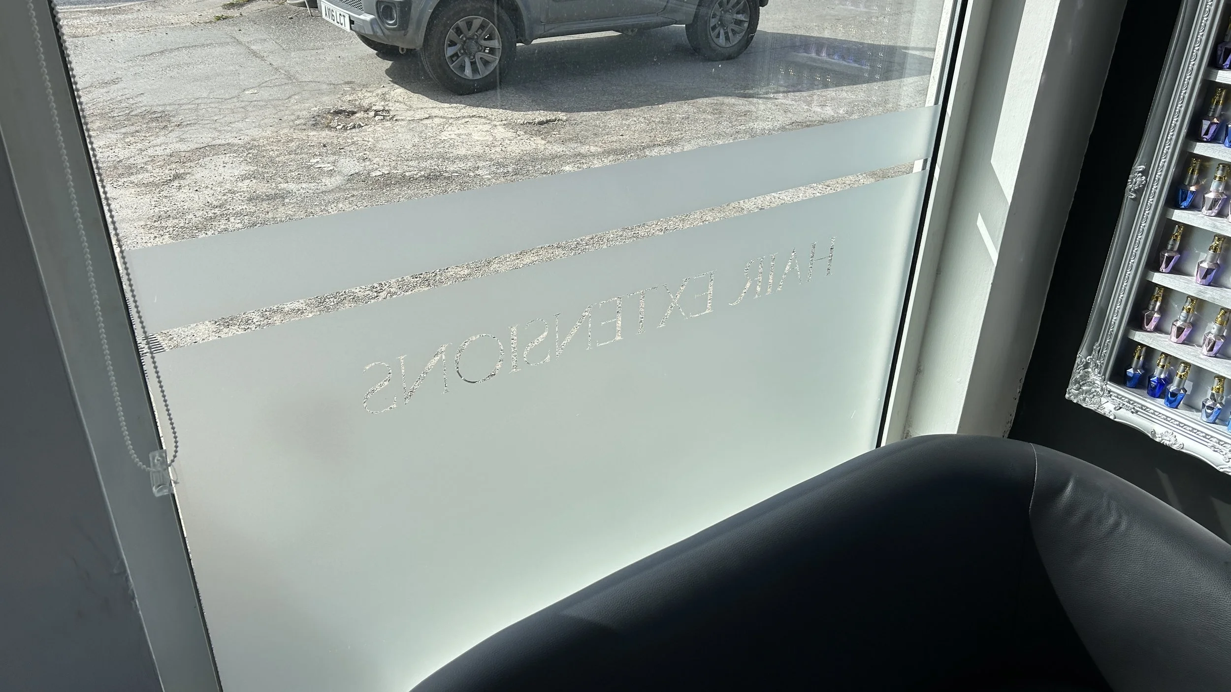 View through a glass door showing a parking area with a black vehicle and a small display of perfume bottles inside a store.