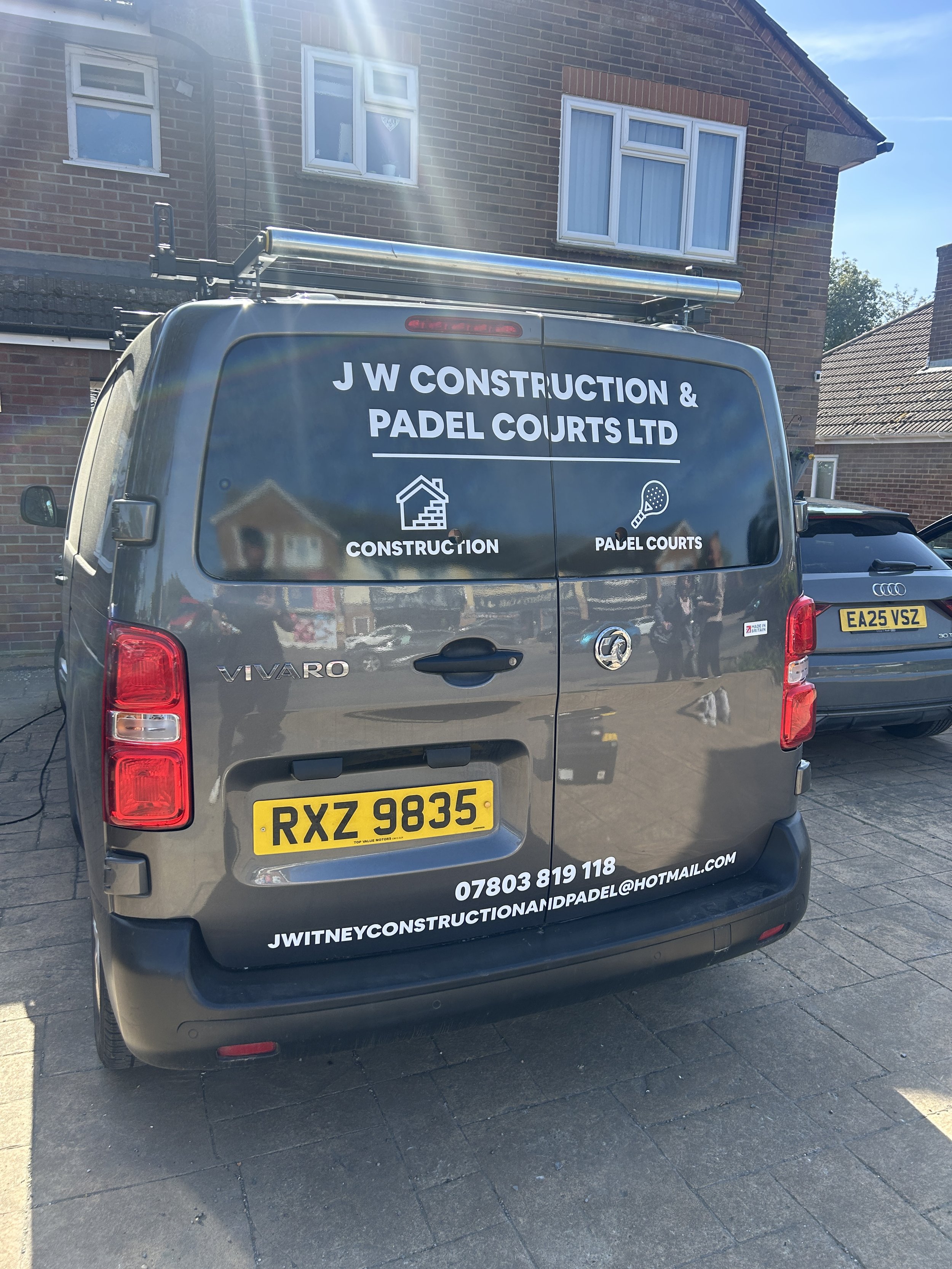 Black van with company name JW Construction & Padel Courts LTD printed on the back, parked on a driveway in front of a brick house. The van has contact details including a phone number, email, and website.