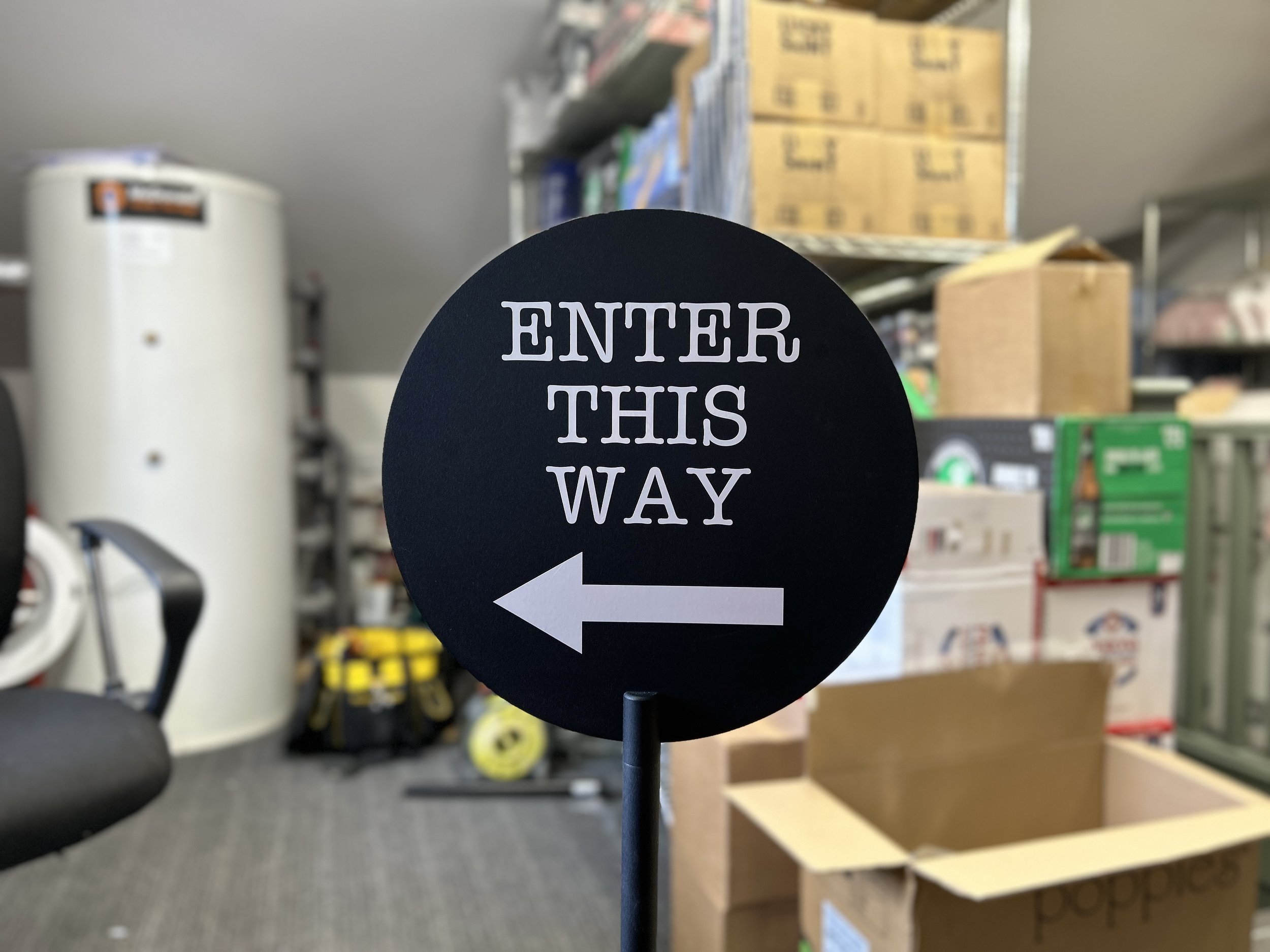 A black circular sign with white text and a white arrow pointing left that reads 'ENTER THIS WAY' in a store aisle.