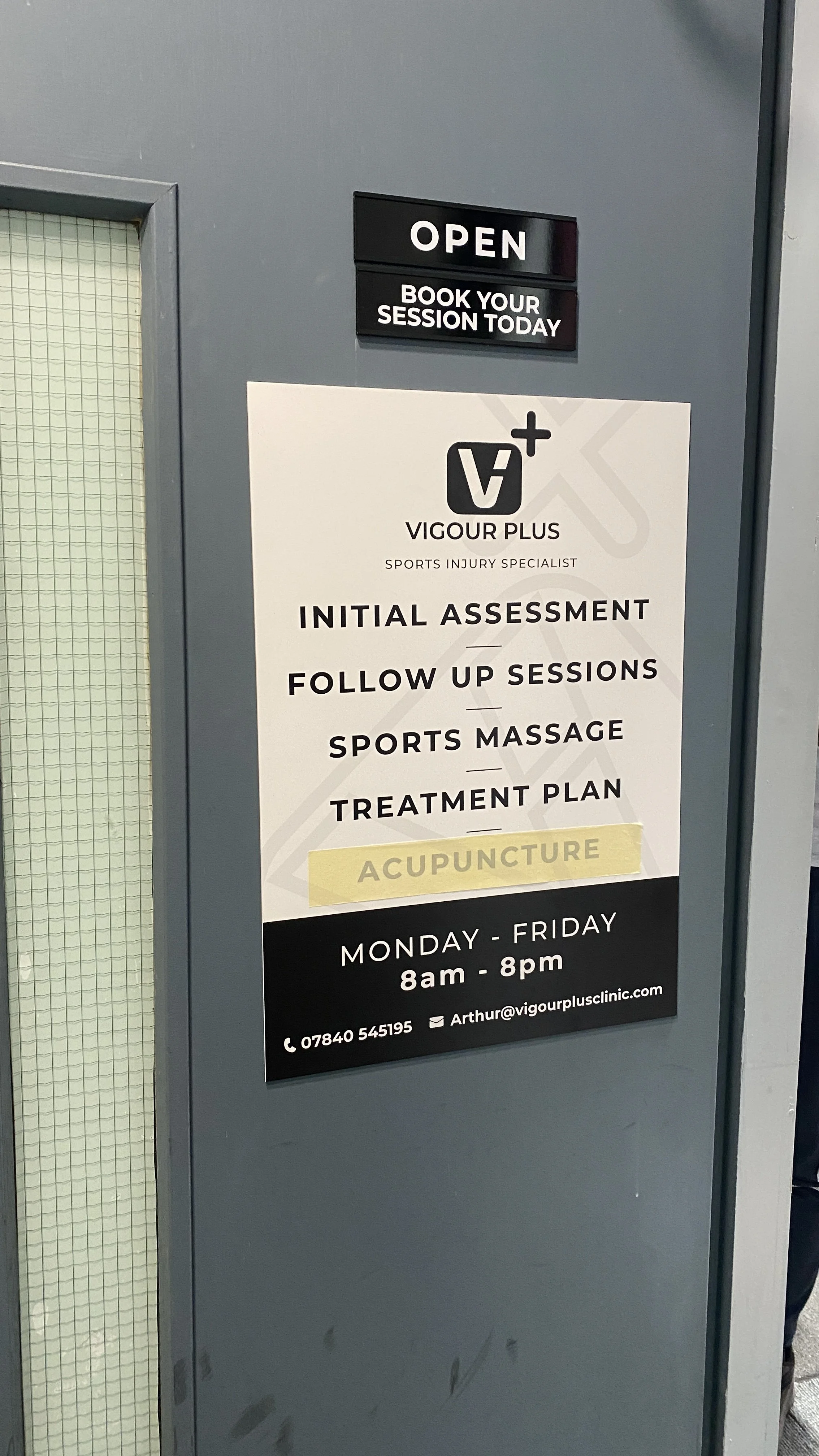 Signboard outside a clinic advertising Vigour Plus sports injury specialist services, including initial assessment, follow-up sessions, sports massage, and acupuncture, with operational hours from Monday to Friday, 8 am to 8 pm.