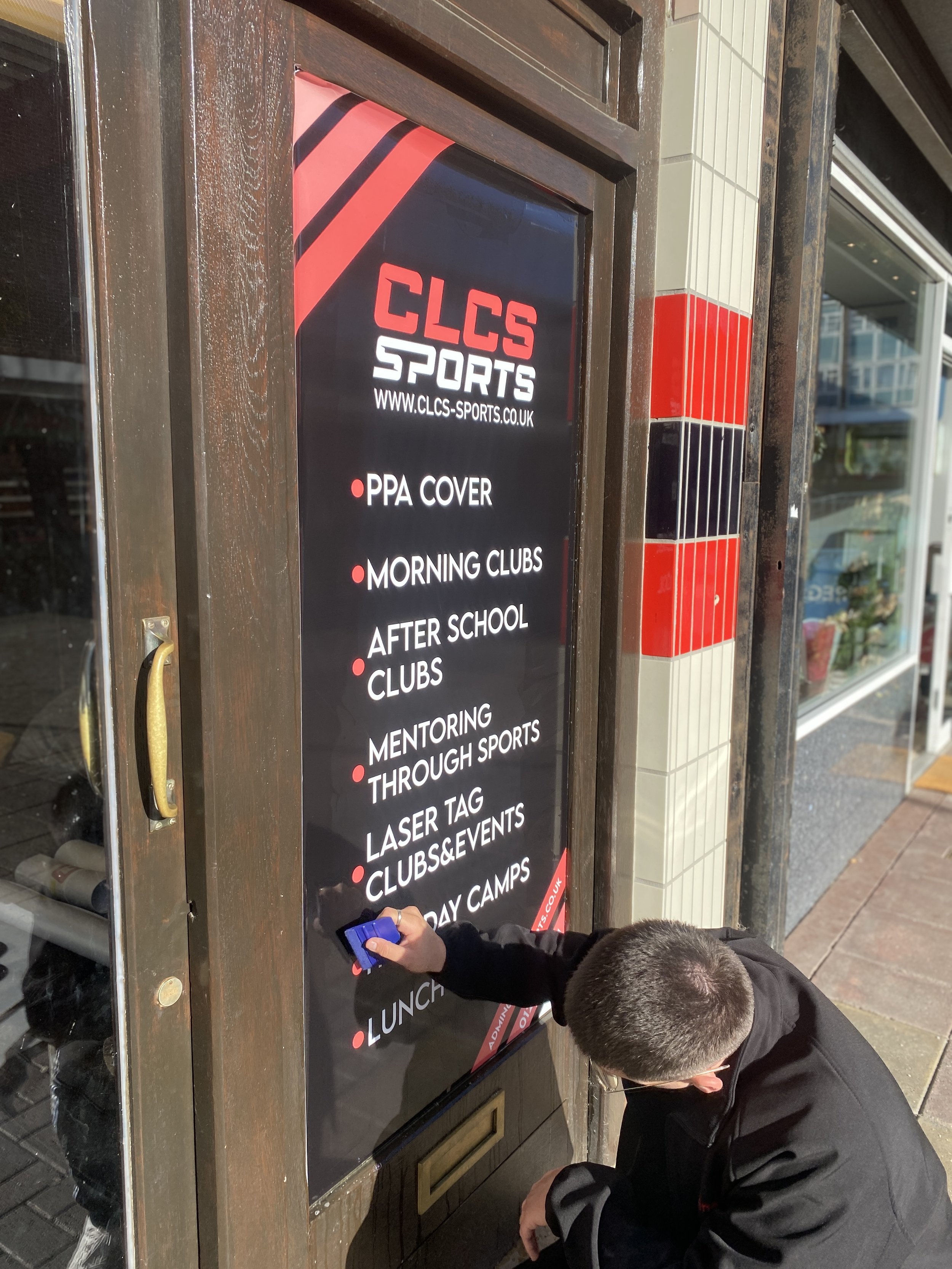 A man cleaning or adjusting a large black sign with red and white text outside a sports shop, listing services like PPA cover, morning clubs, after school clubs, mentoring through sports, laser tag, clubs and events, and holiday camps.
