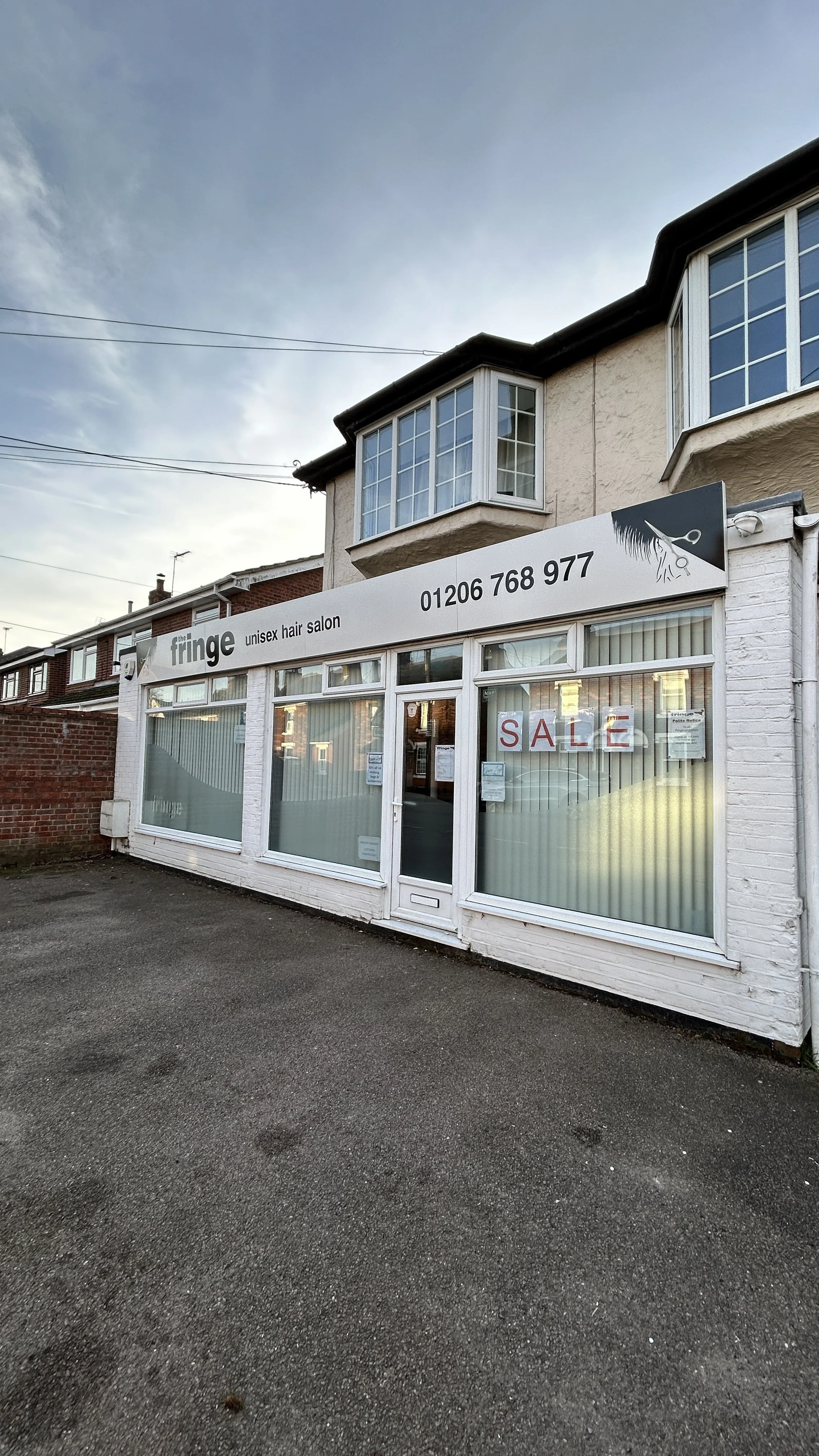 Front view of a white, single-story unisex hair salon with large glass windows and a sale sign in the window. The salon has a sign on the roof with the salon's name, logo, phone number, and description.
