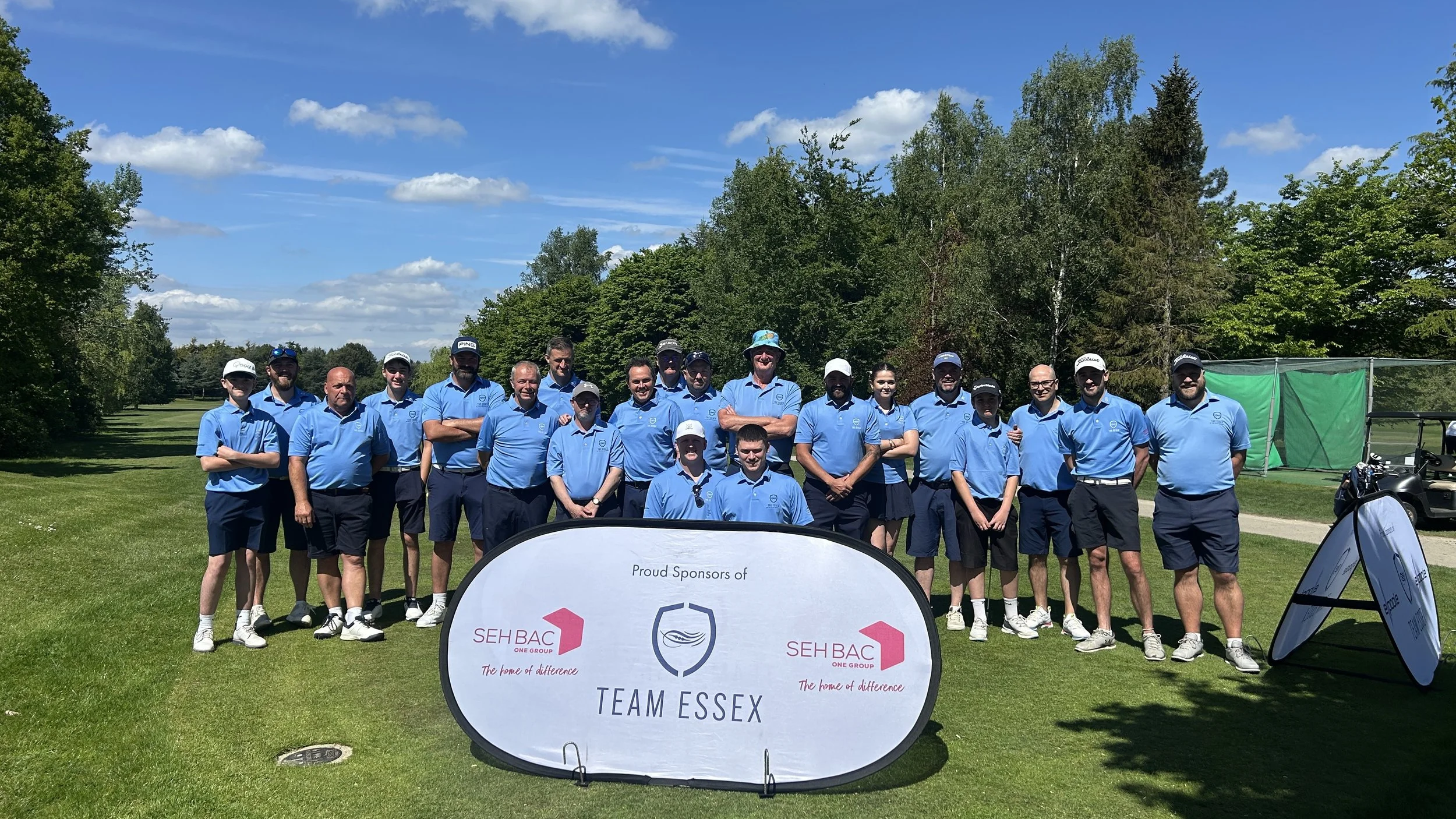 Group of people in blue shirts standing on a golf course, posing for a photo with trees and a partly cloudy sky in the background. There is a sign in front that reads "TEAM ESSEX" and features logos for SEH BAC and another organization.