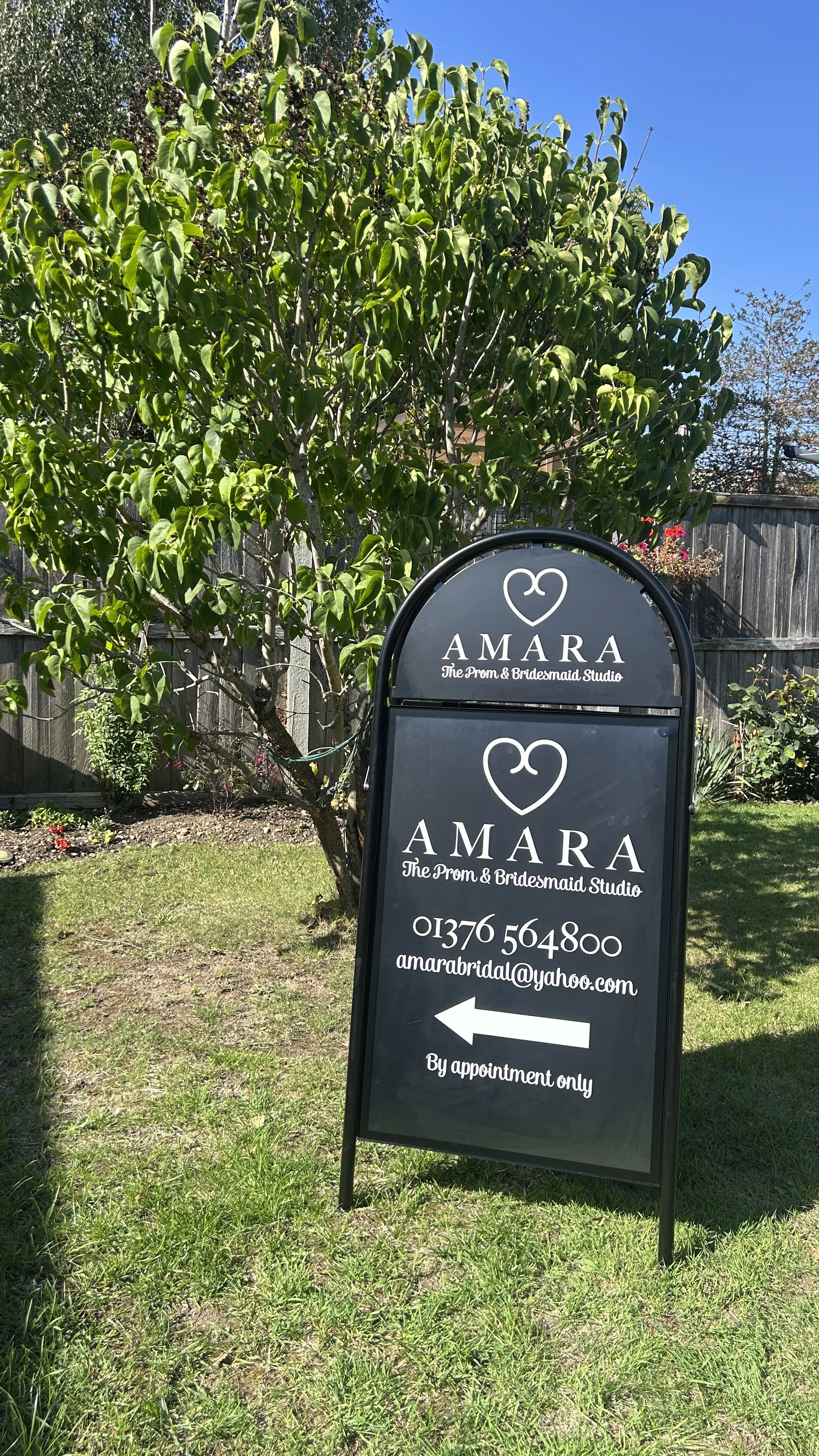 A black signboard with white text advertising "AMARA The Prom & Bridesmaid Studio," an arrow pointing left, and a phone number and email address, placed on a grassy area beside a tree and wooden fence under a bright blue sky.