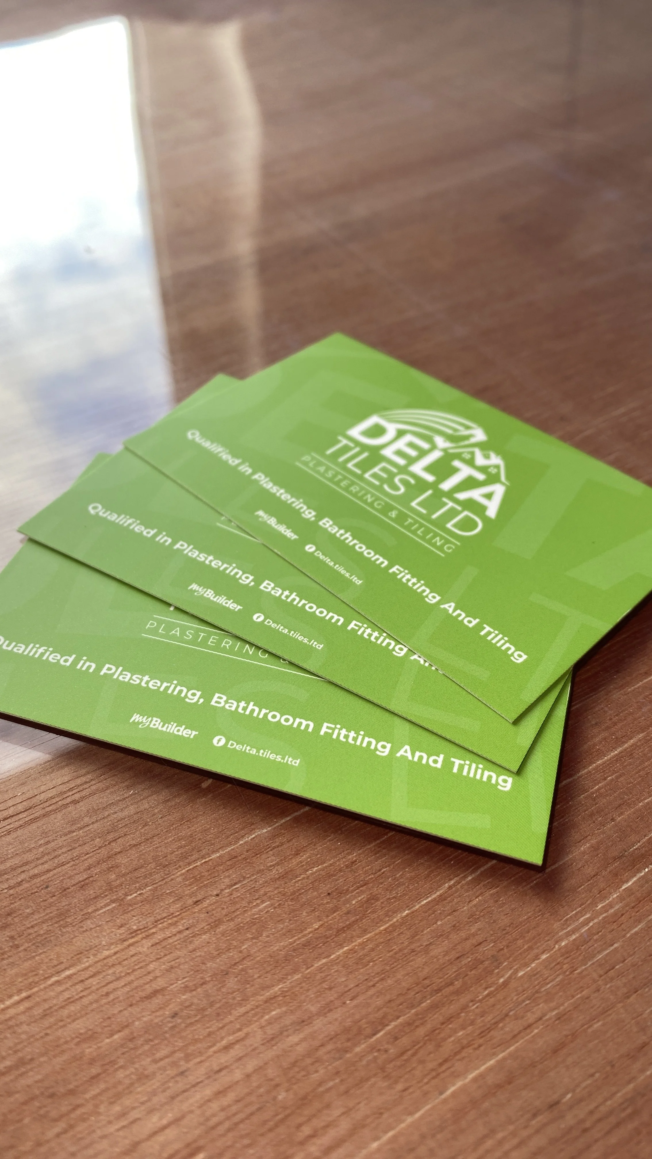Business cards for Delta Tiles Ltd, specializing in plastering, bathroom fitting, and tiling, placed on a wooden surface.