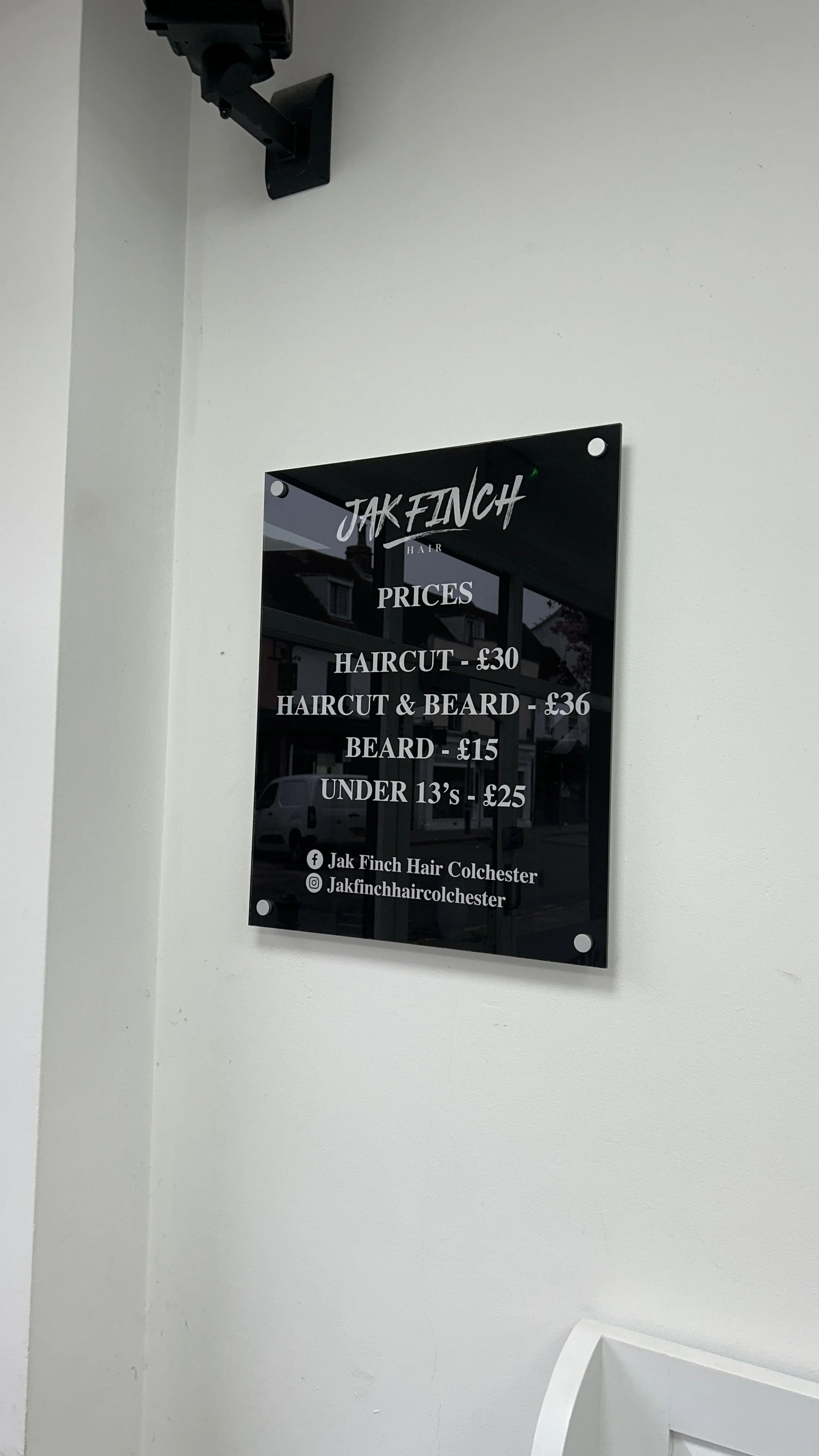 A black sign with white text listing hair salon prices, including haircut for 30 pounds, haircut and beard for 36 pounds, beard for 15 pounds, and haircuts under 13 years old for 25 pounds, displayed on a white wall.