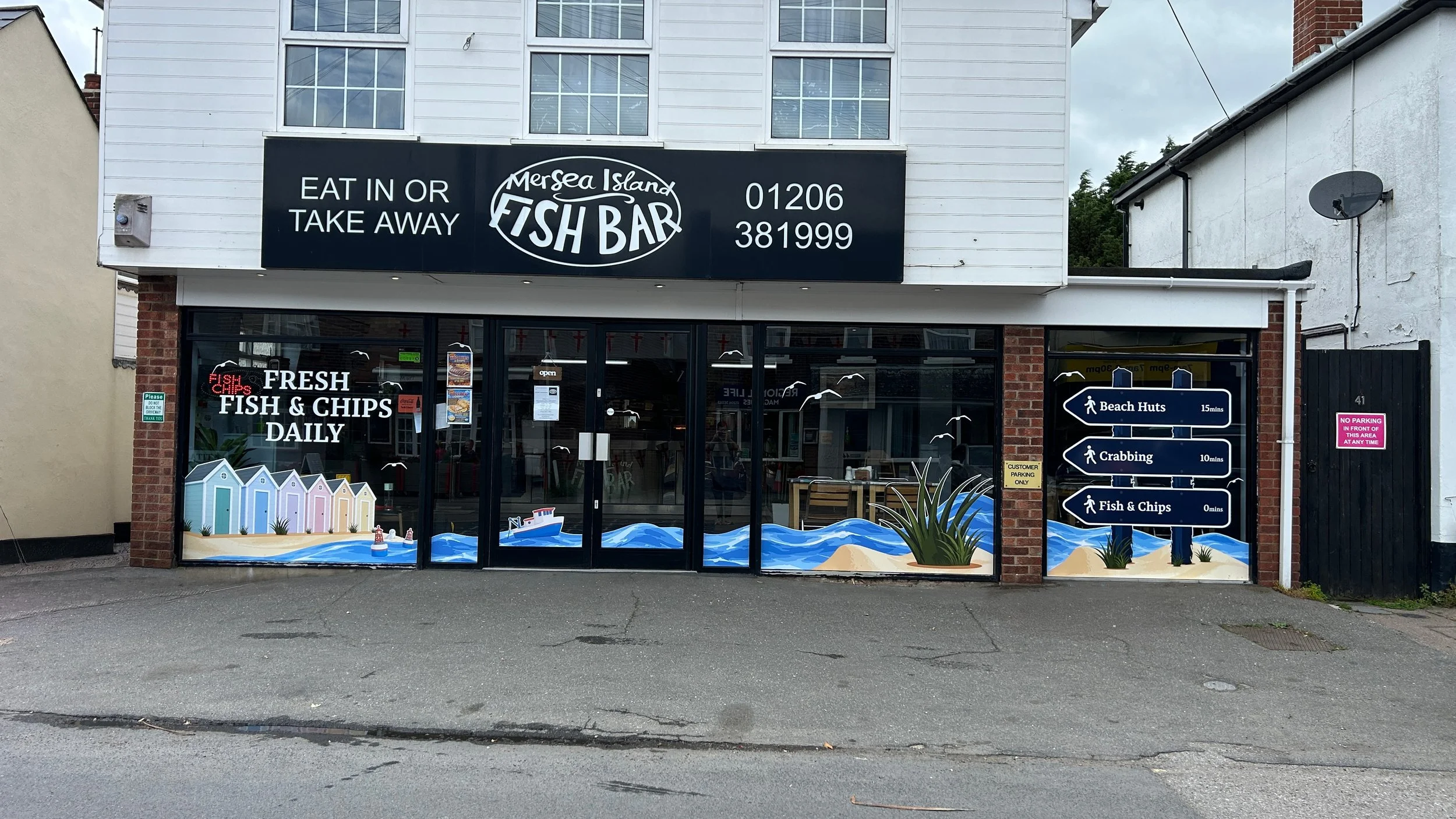 Front view of a seafood restaurant called 'Mersea Island Fish Bar' with a black signboard displaying contact number and service options for eat-in or take-away. The shopfront has a large glass window with colorful beach-themed decorations, and a sign