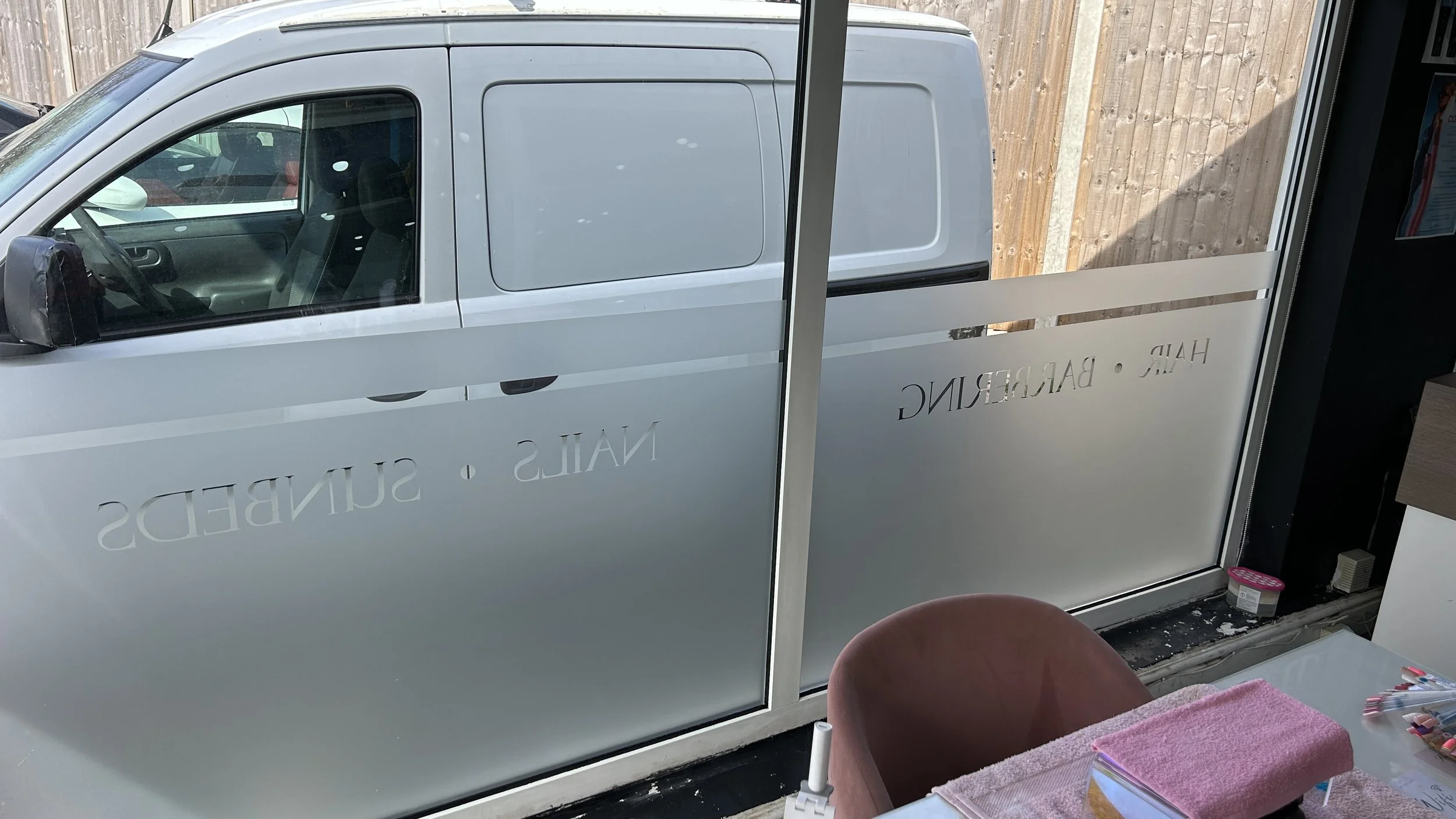 View from inside a nail salon looking out a glass window at a parked white van. The window has the words 'NAILS' and 'SALON' written on it. Inside, there is a pink towel on a table, a small container of nail tools, and a pink cushioned chair.