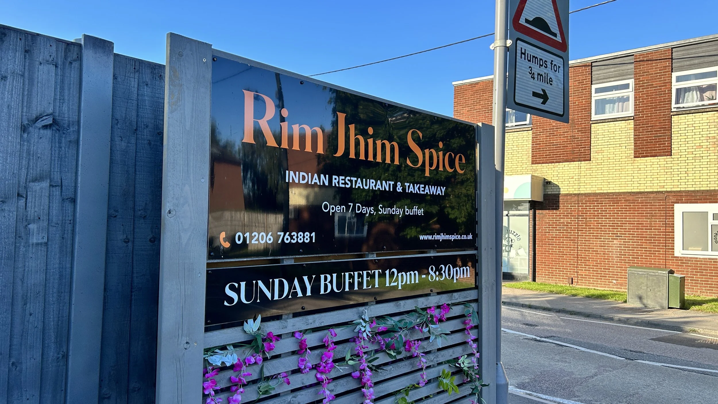 Signboard for Rim Jhimu Spice, an Indian restaurant and takeaway, with a Sunday buffet from 12 pm to 8:30 pm, and a small sign indicating humps for 3.4 miles. The background shows a brick building and a clear blue sky.