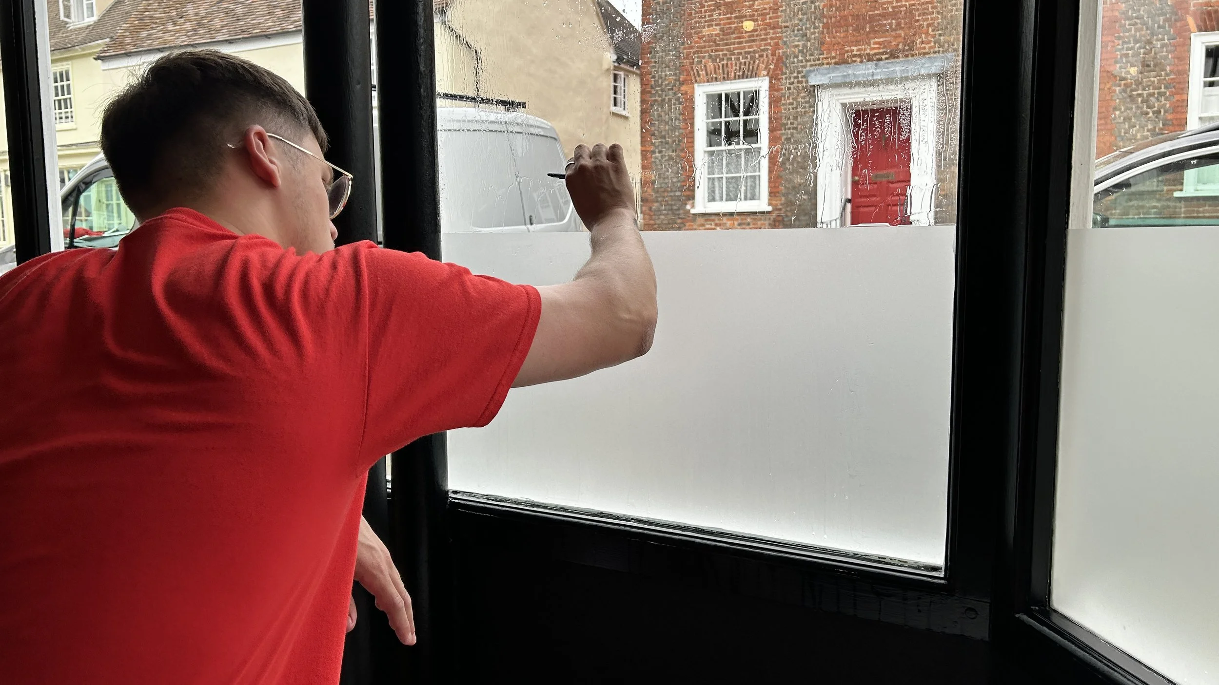 A young man in a red shirt writing on a window with a marker, viewed from behind inside a shop or café, with buildings visible outside.