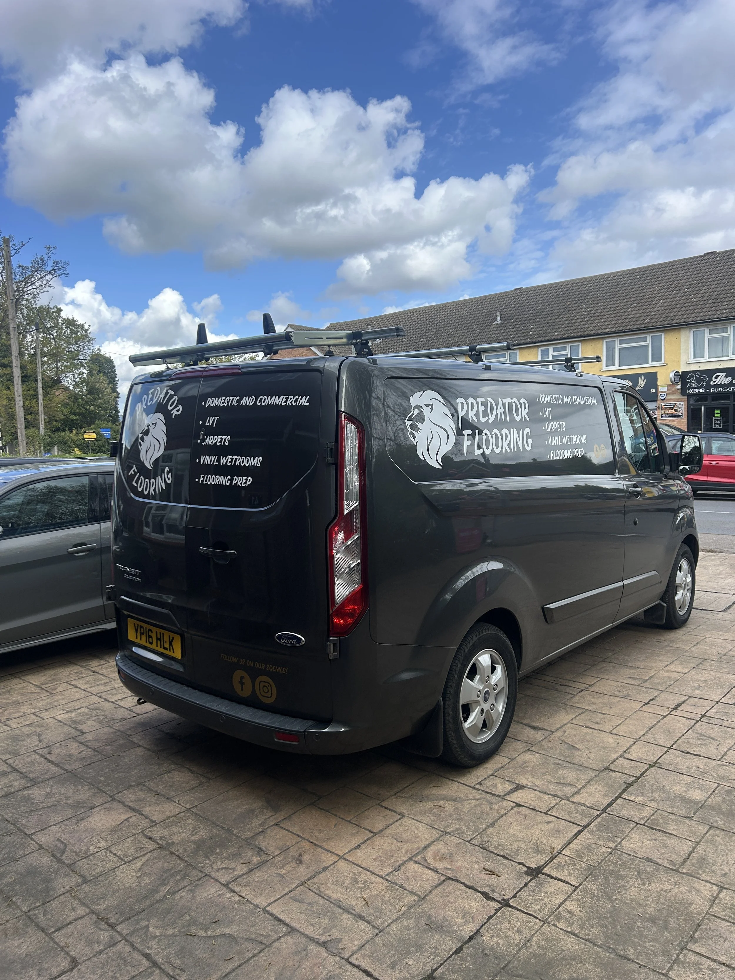 Black van labeled 'Predator Flooring' parked on a brick sidewalk with a row of shops and houses in the background under a partly cloudy sky.