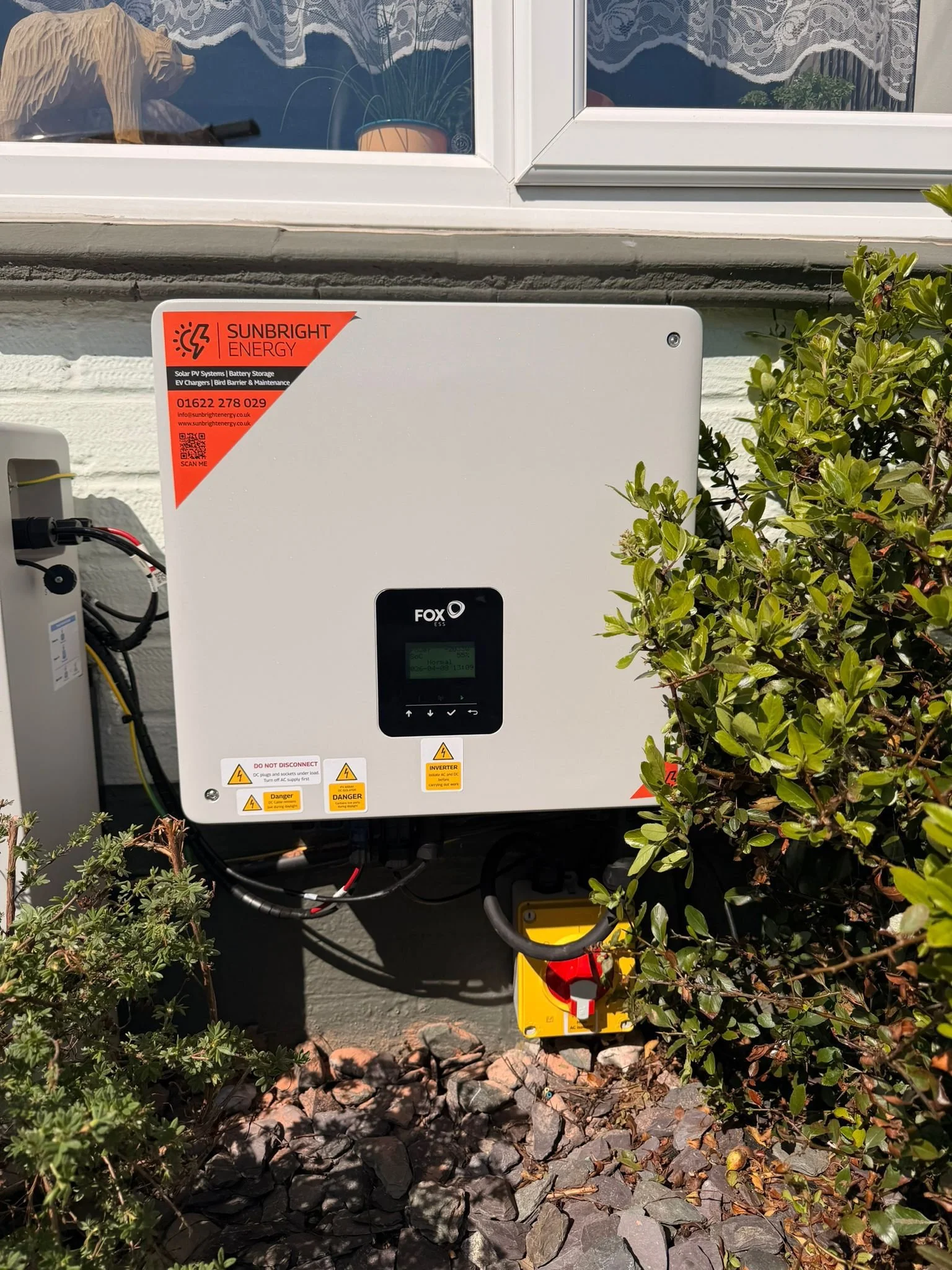 Photograph of a solar power system mounted outside on a wall, partially obscured by shrubbery and rocks. The system has warning labels and a red sticker with the logo 'Sunbright Energy.' A window with lace curtains is visible above the system.