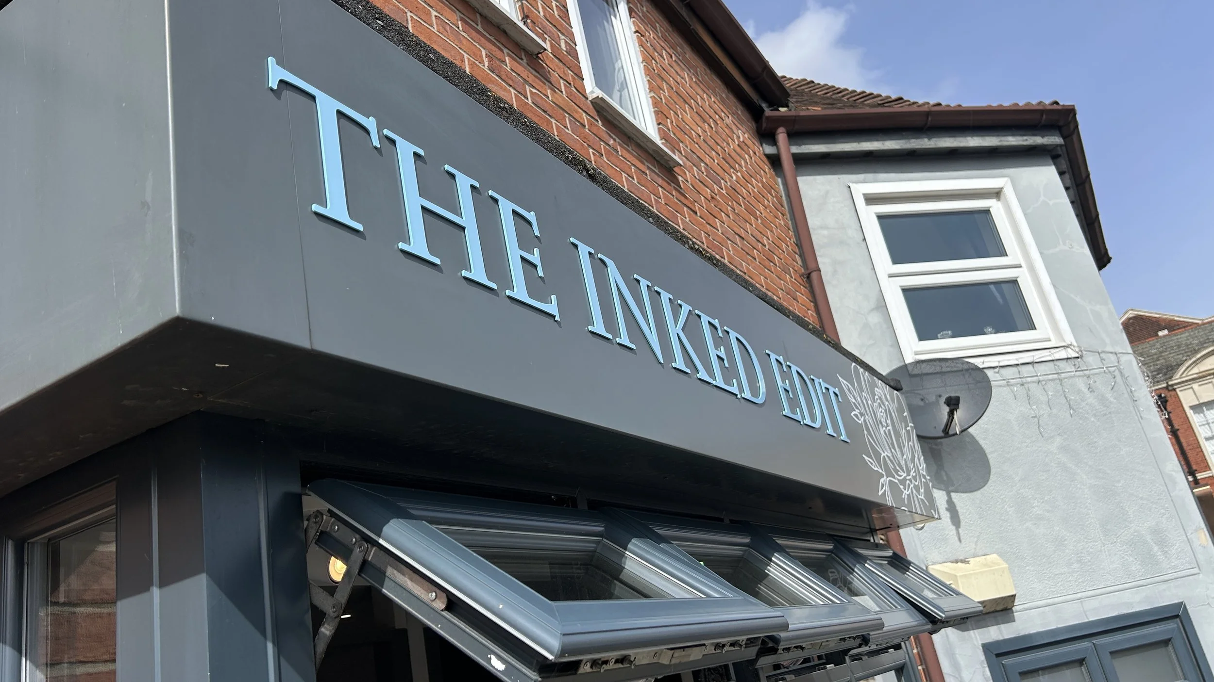 Photo of a storefront with a sign that reads 'The Inked Edit' in light blue letters on a gray background. The storefront is on a building with a red brick upper facade and a gray lower facade featuring trim and a window above.