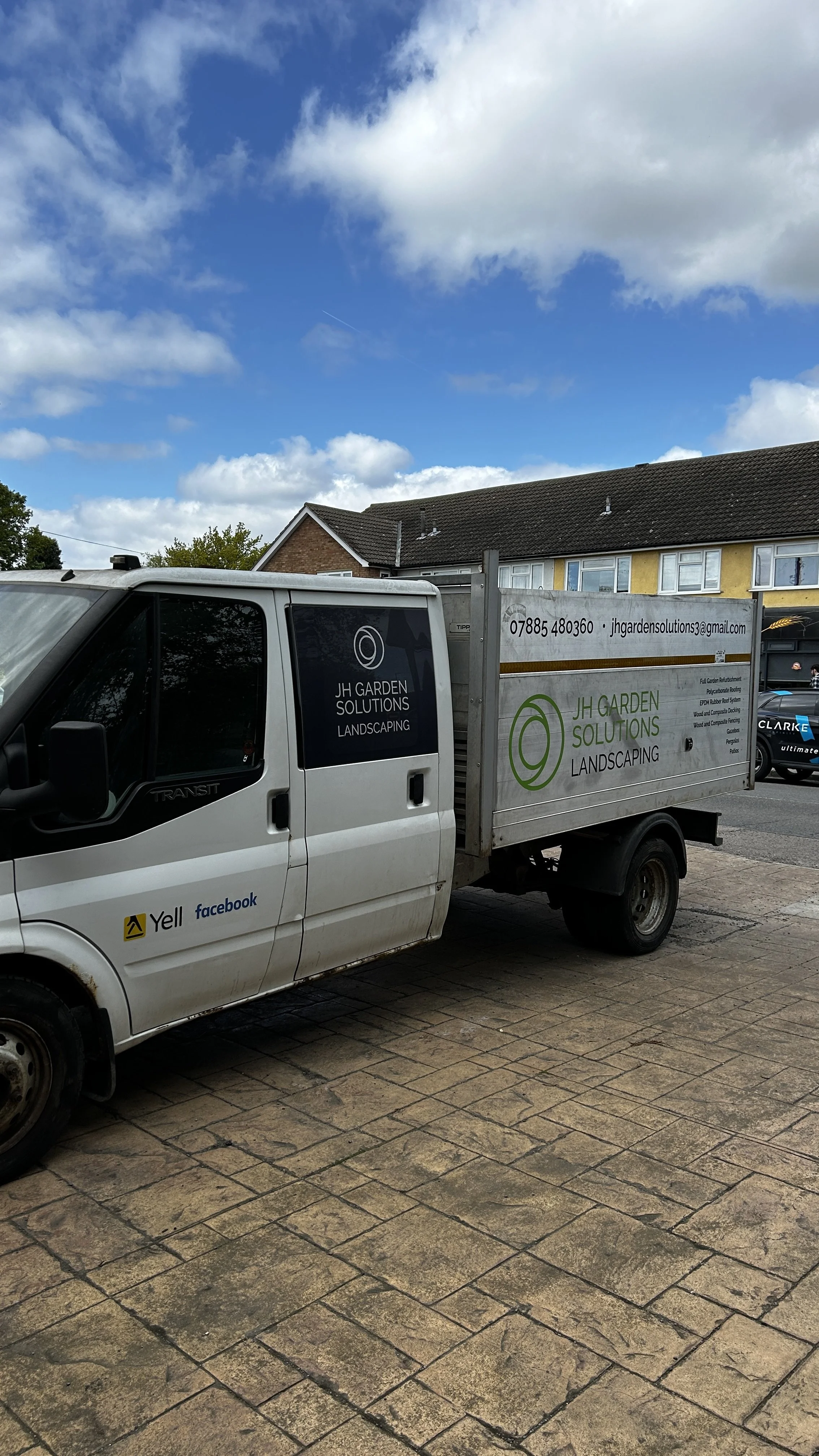 A white utility truck parked on a brick-paved driveway with signs advertising JH Garden Solutions Landscaping. The truck's side panel displays contact information, and the door shows Facebook and Yelp logos. In the background are houses with pitched 