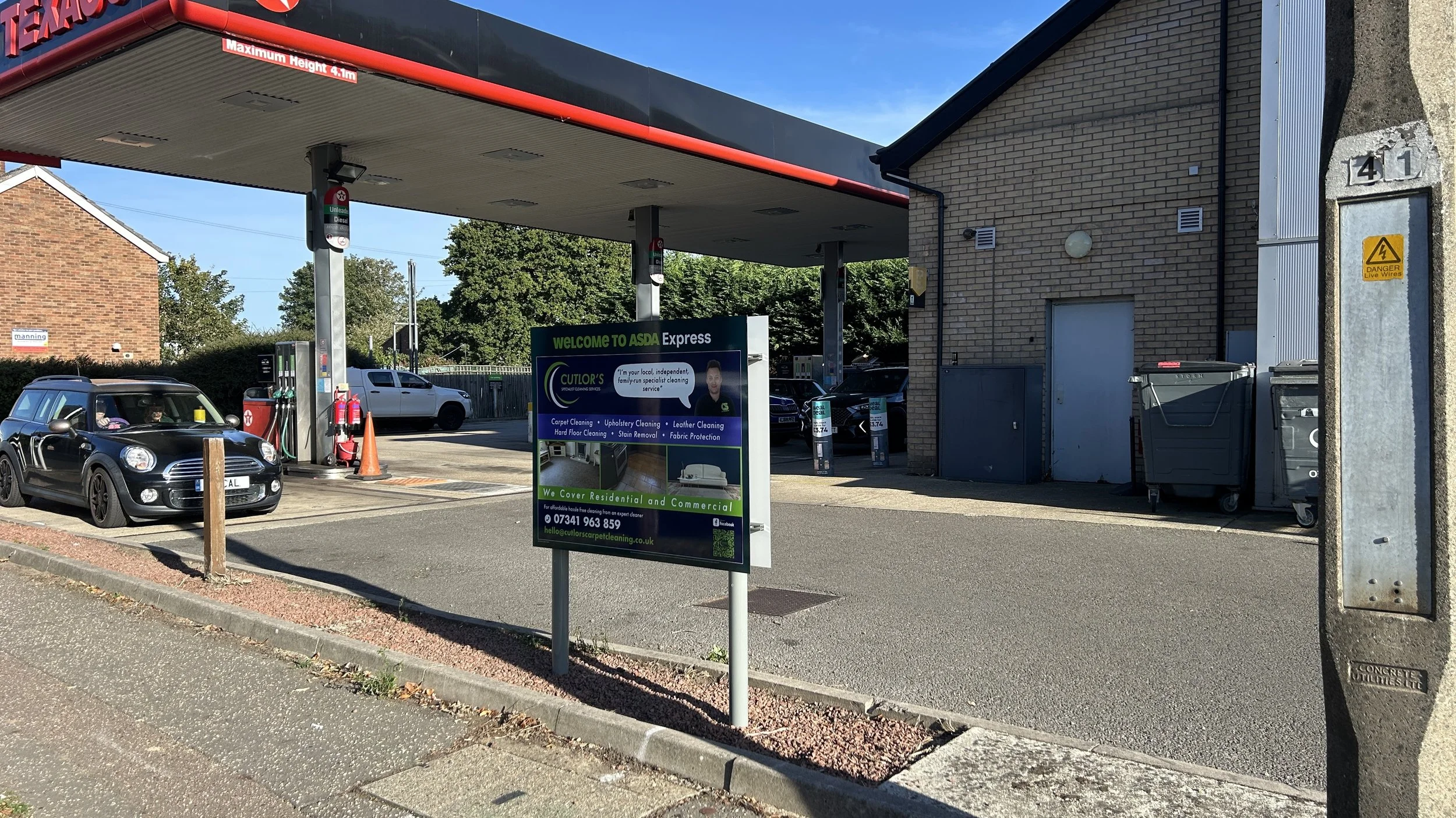 A car wash and cleaning service outside a building with a sign advertising carpet, upholstery, and leather cleaning. There are several cars parked near the area, including a black Mini Cooper. The building has a brick exterior with a garage door and 