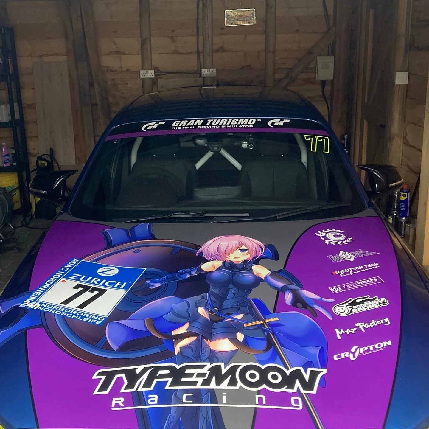 A race car with a colorful anime-style character graphic on the hood, featuring a female character with pink hair and blue eyes, wearing a blue and black outfit, with various sponsor logos including TZYYMOON RACING, Zurich, and others, inside a garag
