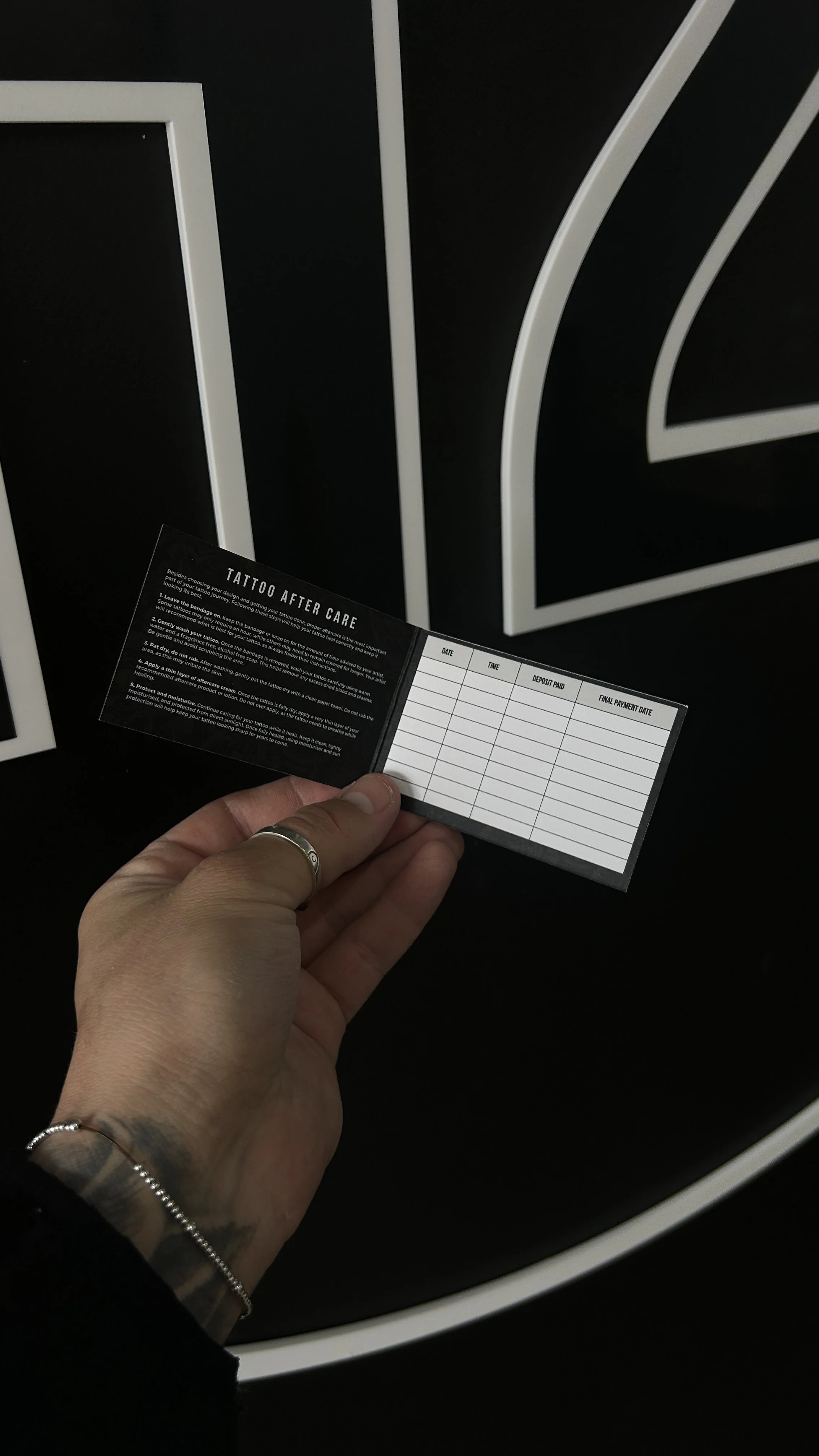 A hand holding a tattoo aftercare instruction card with sections for date, time, deposit paid, and final payment date, against a black background with white lines.